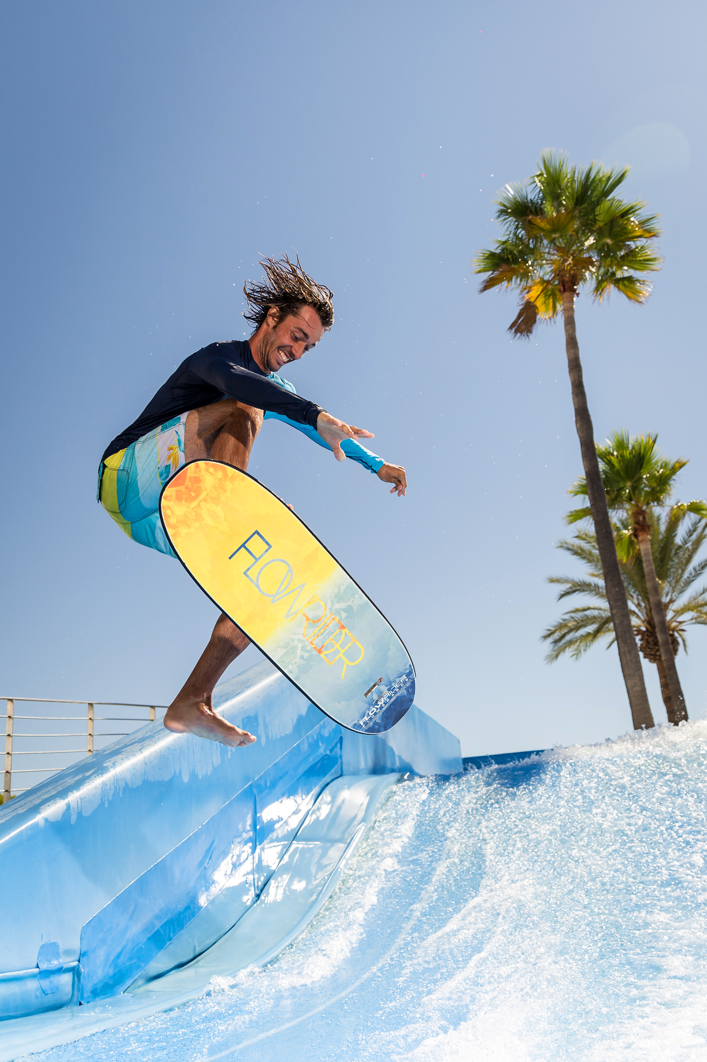 Surfer jumping on Flowrider wave simulator, palm trees, blue sky.
