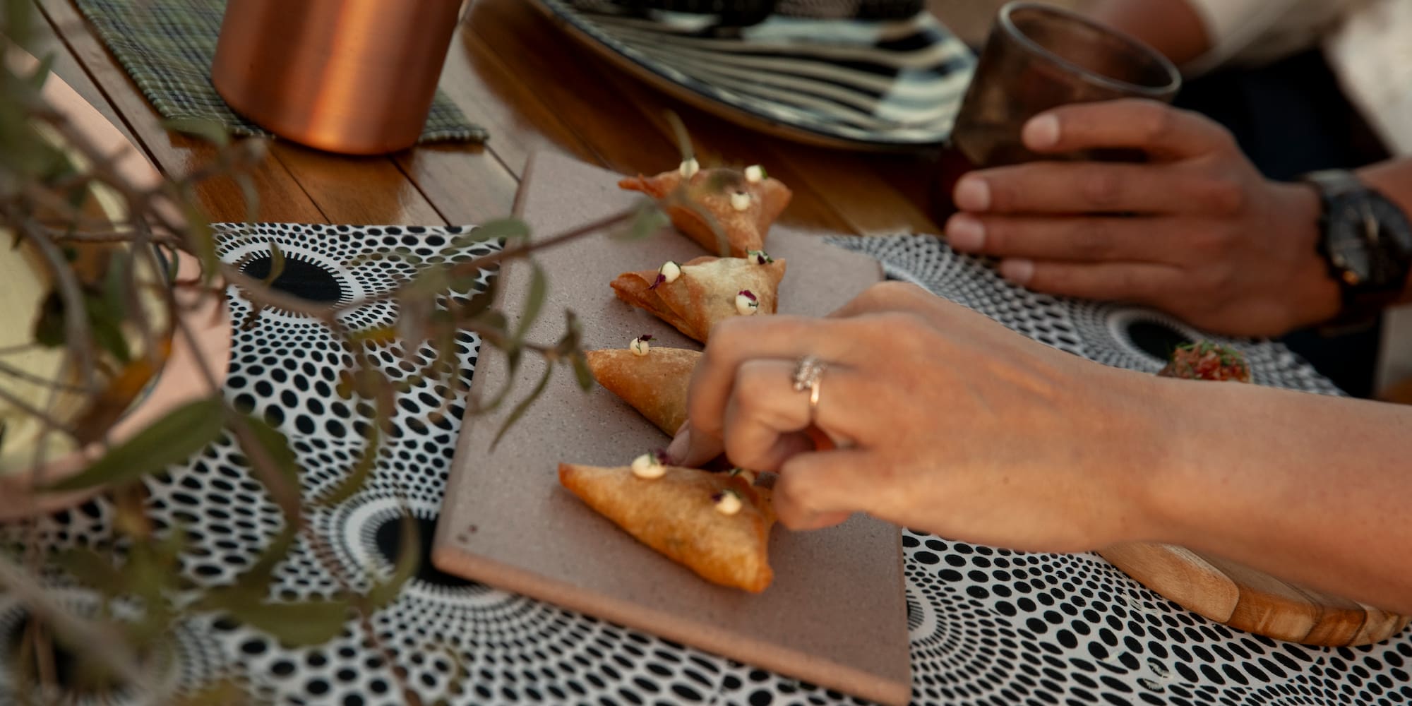 Hand grabs a fried triangular samosa from a board on a patterned dining table.
