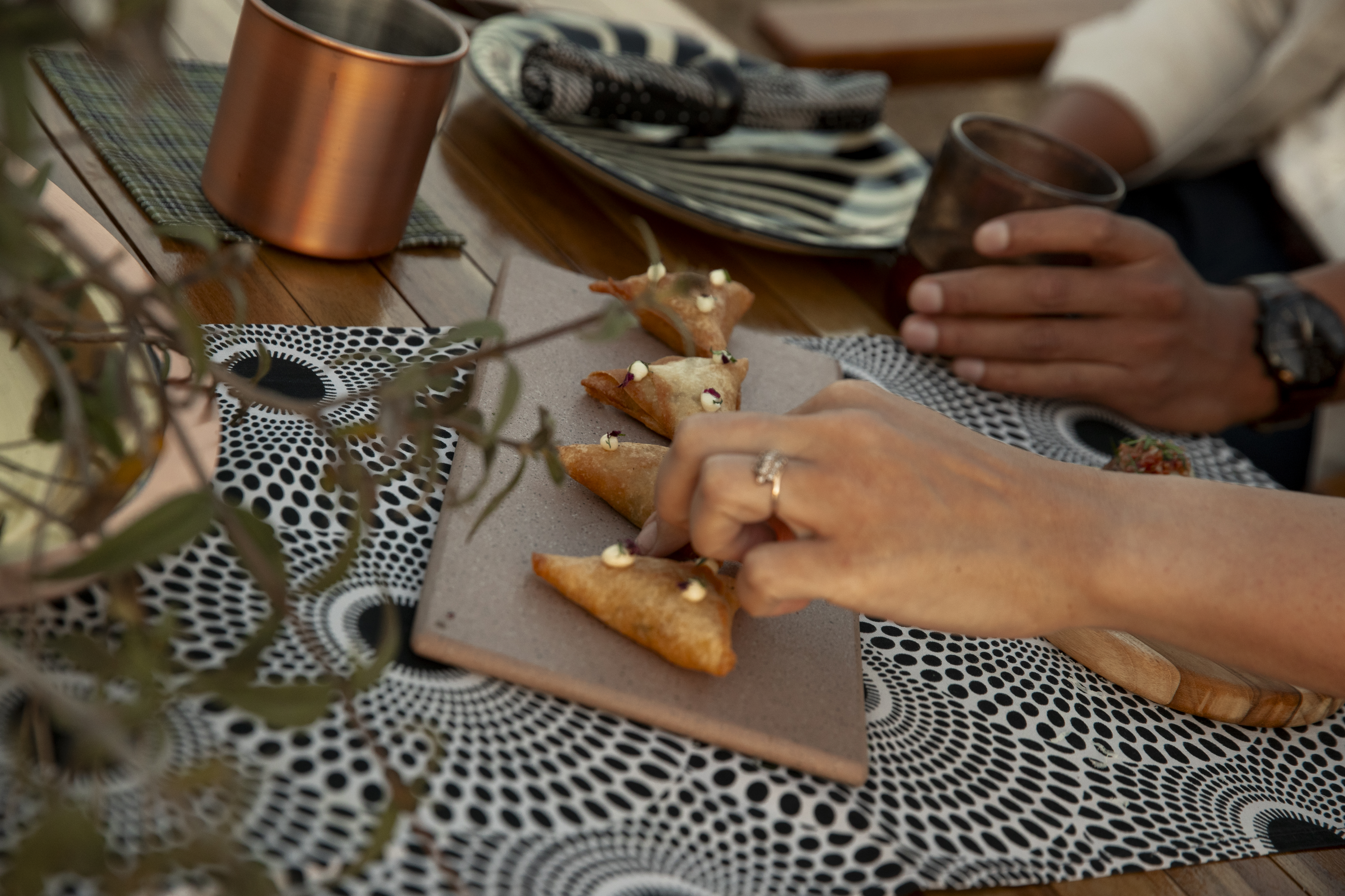 Hand grabs a fried triangular samosa from a board on a patterned dining table.