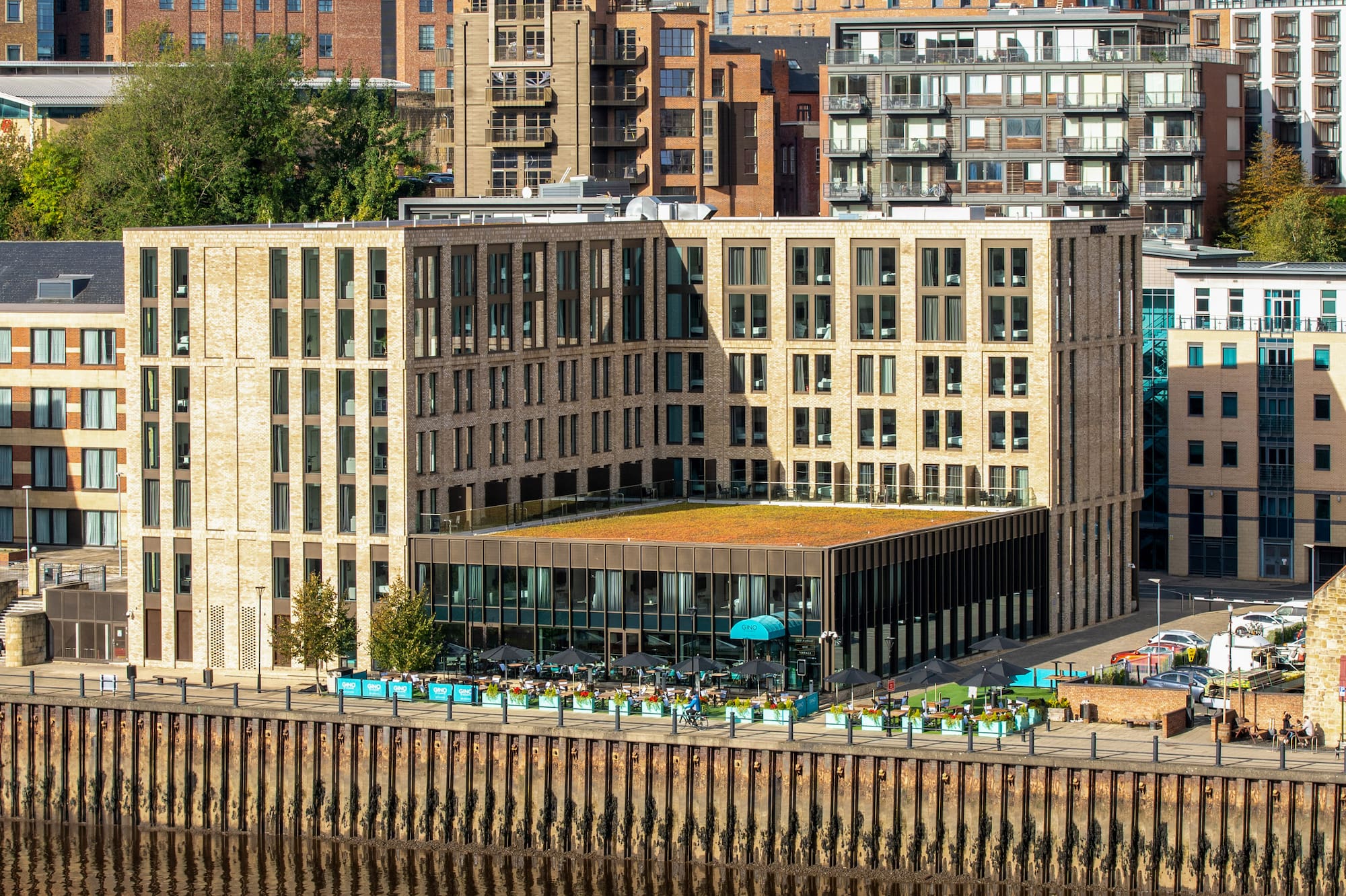 Modern riverfront development with restaurant, outdoor dining, and urban skyline.