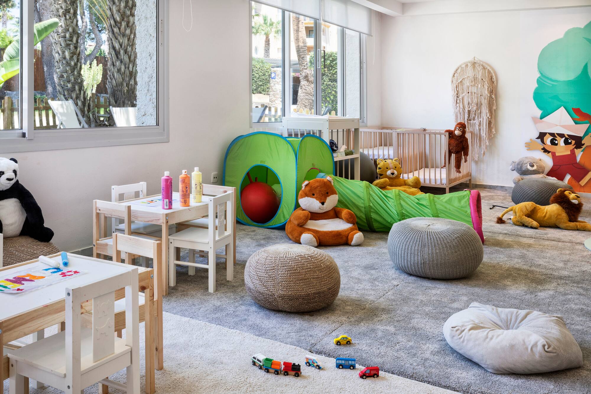 Bright kids playroom with play tent, cribs, art tables, and stuffed animals.