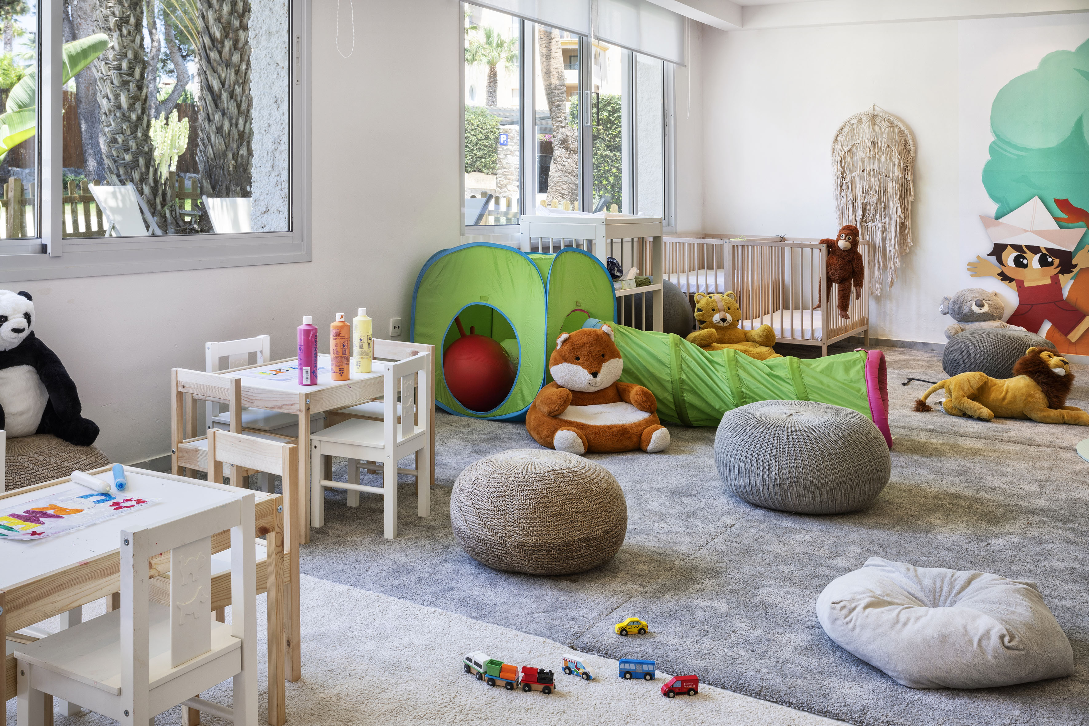 Bright kids playroom with play tent, cribs, art tables, and stuffed animals.