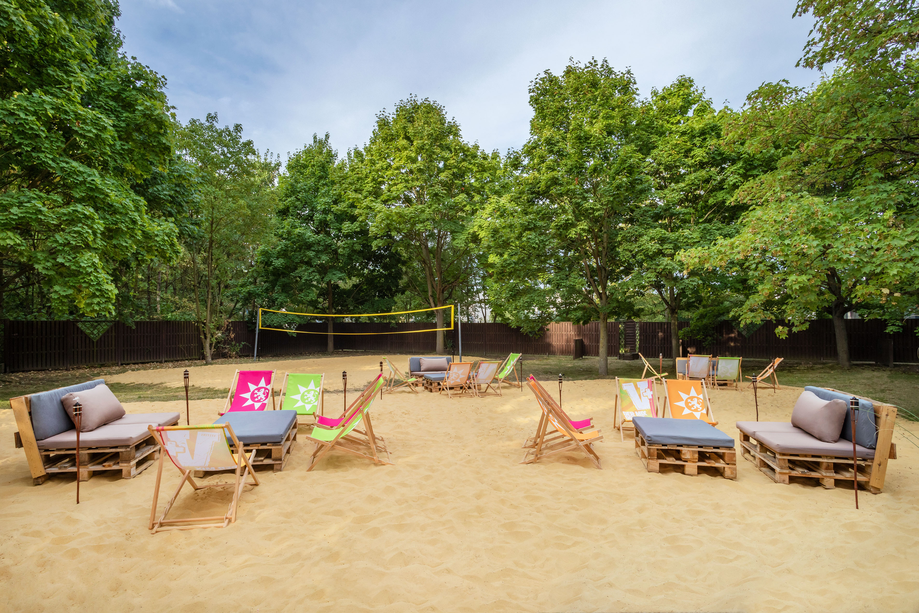 Outdoor sand volleyball court with lounge chairs and pallet furniture under trees.