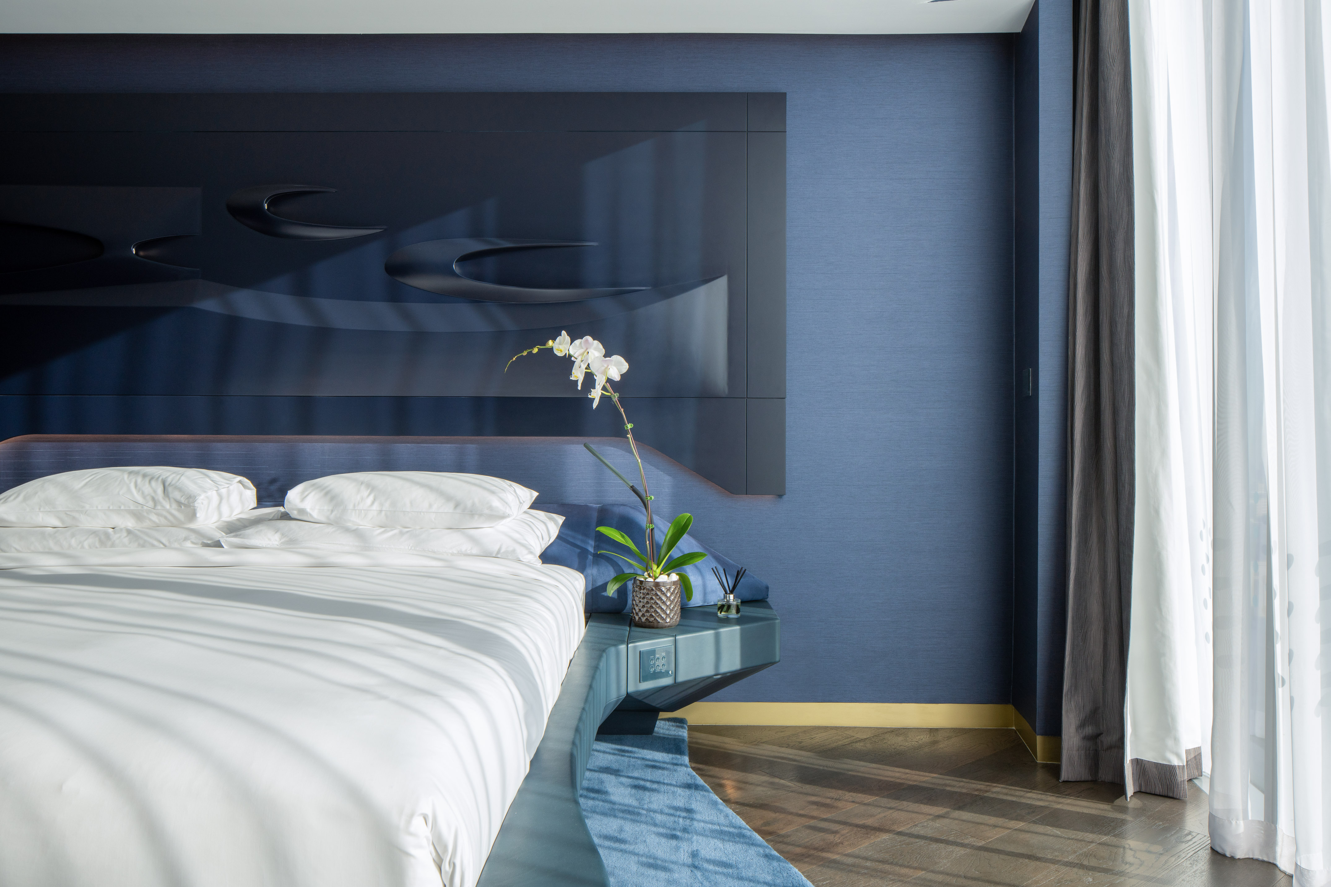 Modern blue bedroom with white bed, orchid, and sculptural headboard.