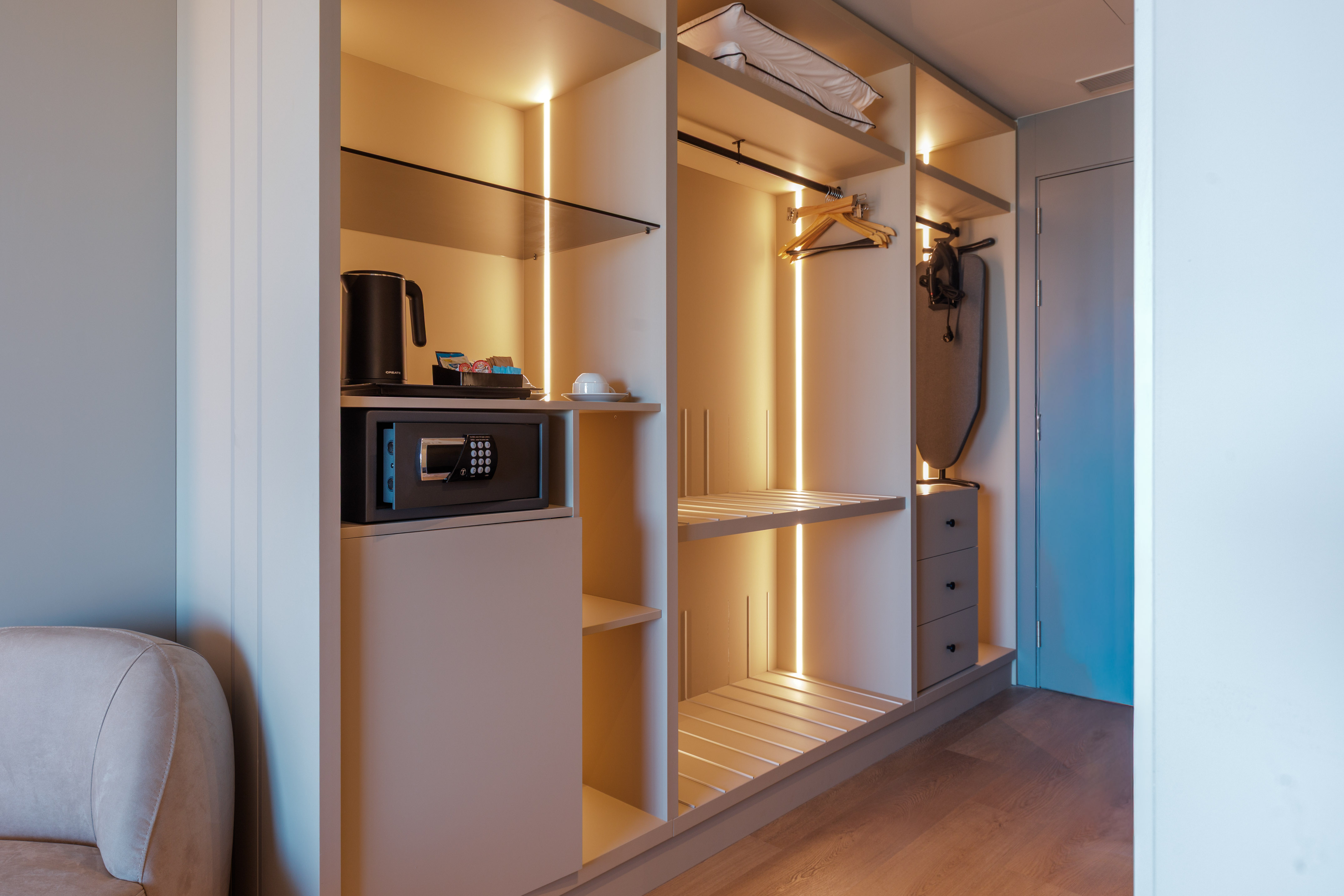 Modern hotel wardrobe with illuminated shelves, safe, kettle, and ironing board.