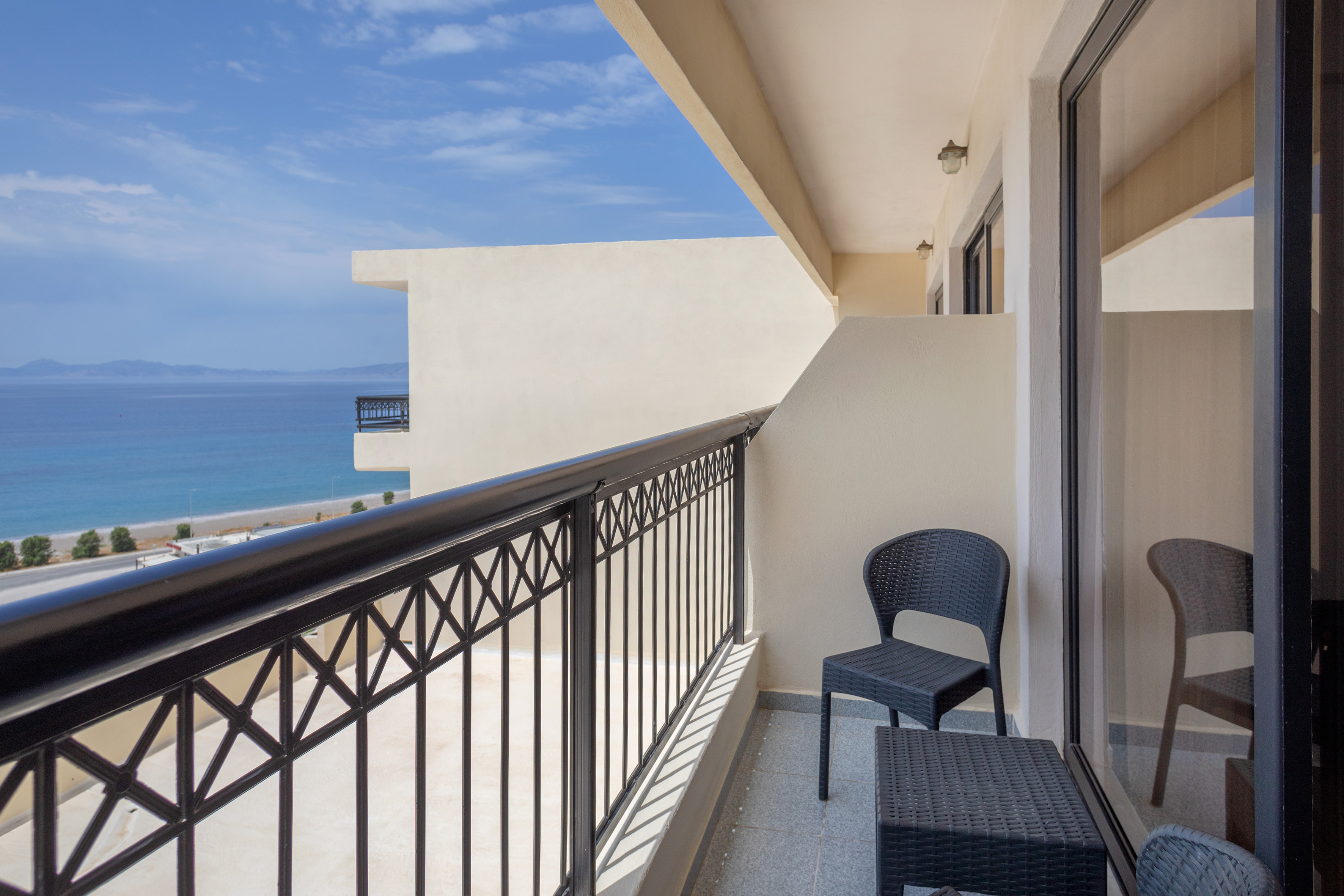 Hotel balcony with sea view, black railing, outdoor seating, beach, blue sky.