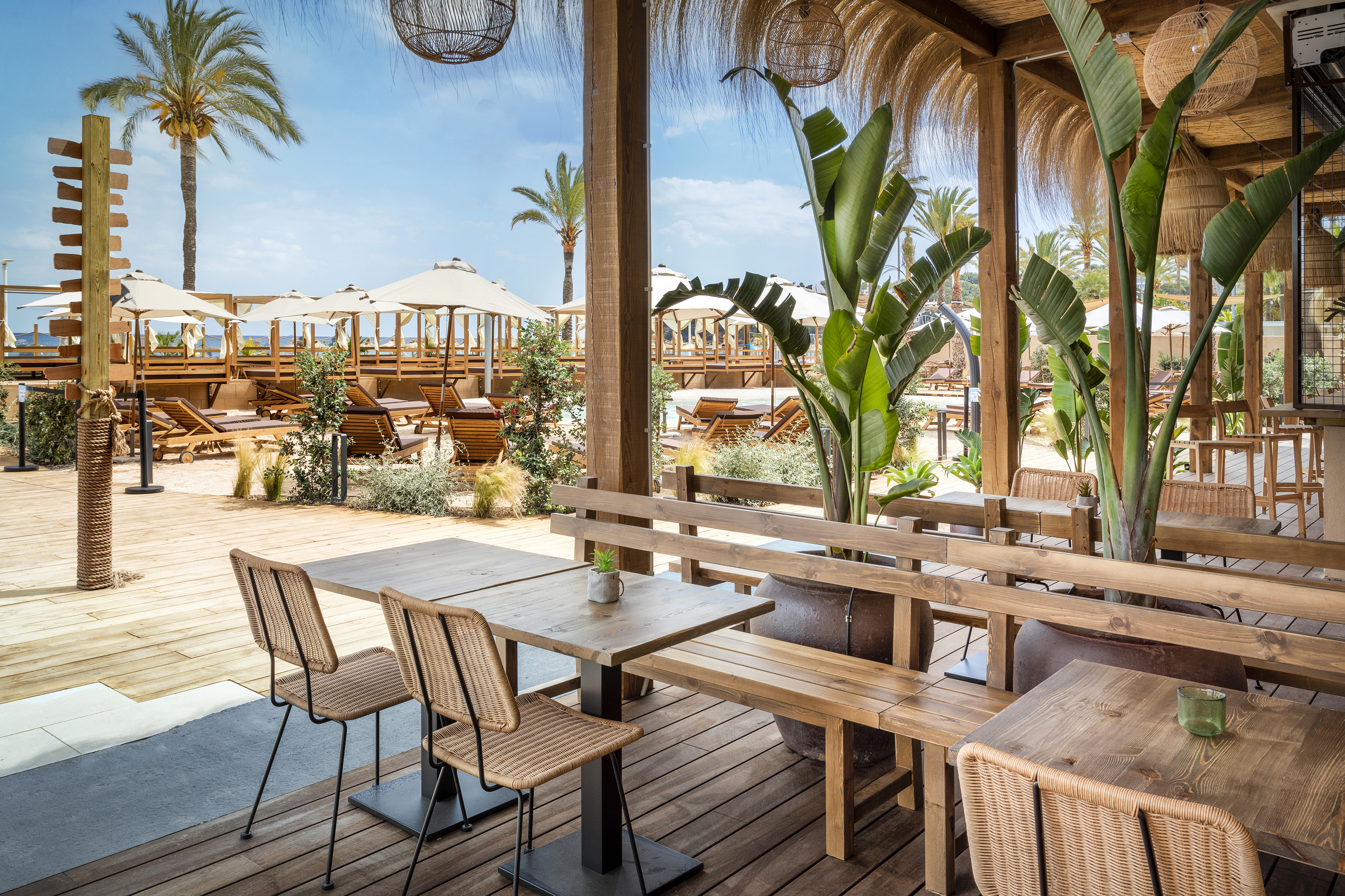Beach club restaurant patio with wooden tables, tropical plants, lounge chairs.