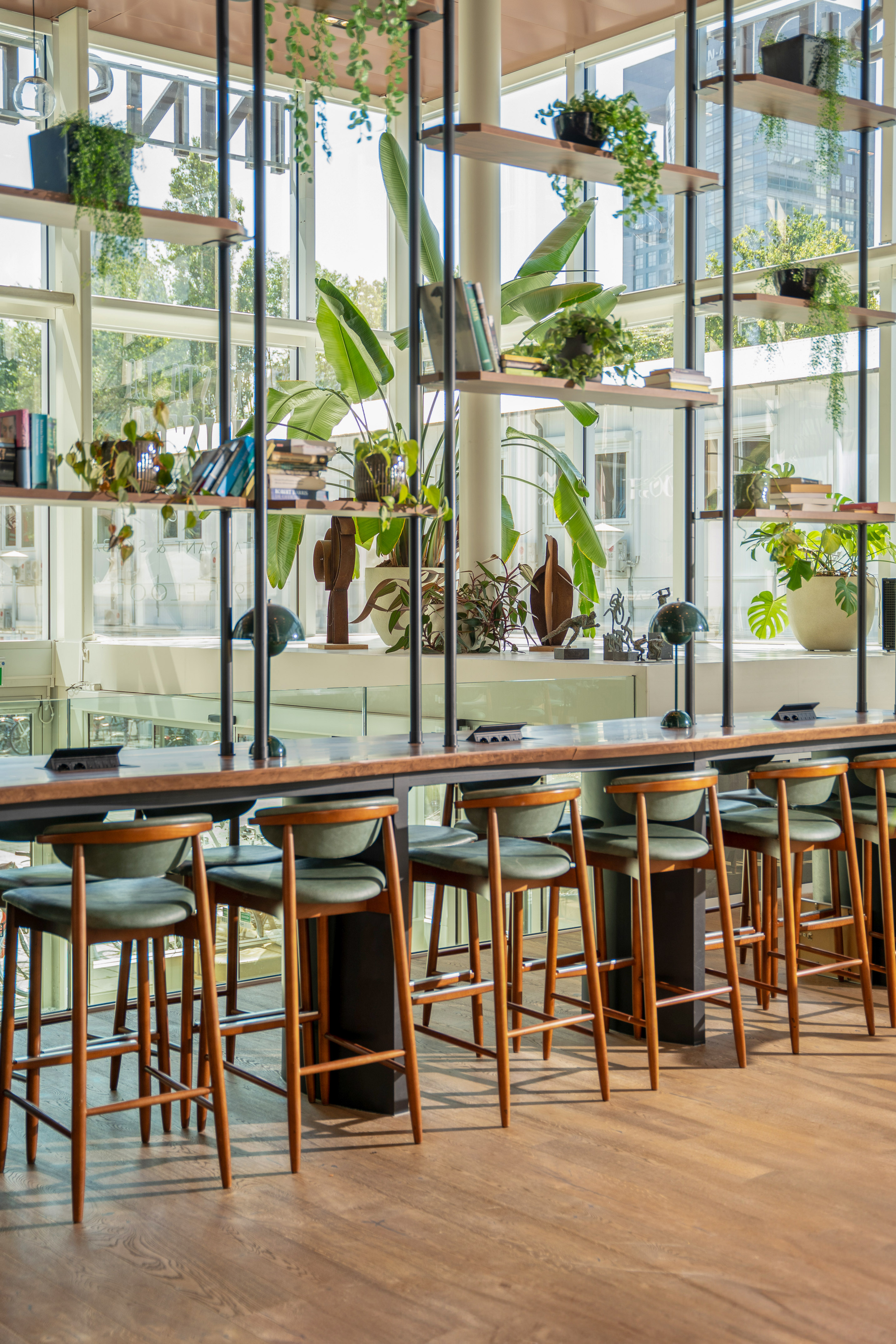 Modern cafe bar with long wooden table, green bar stools, and plant-filled shelves.