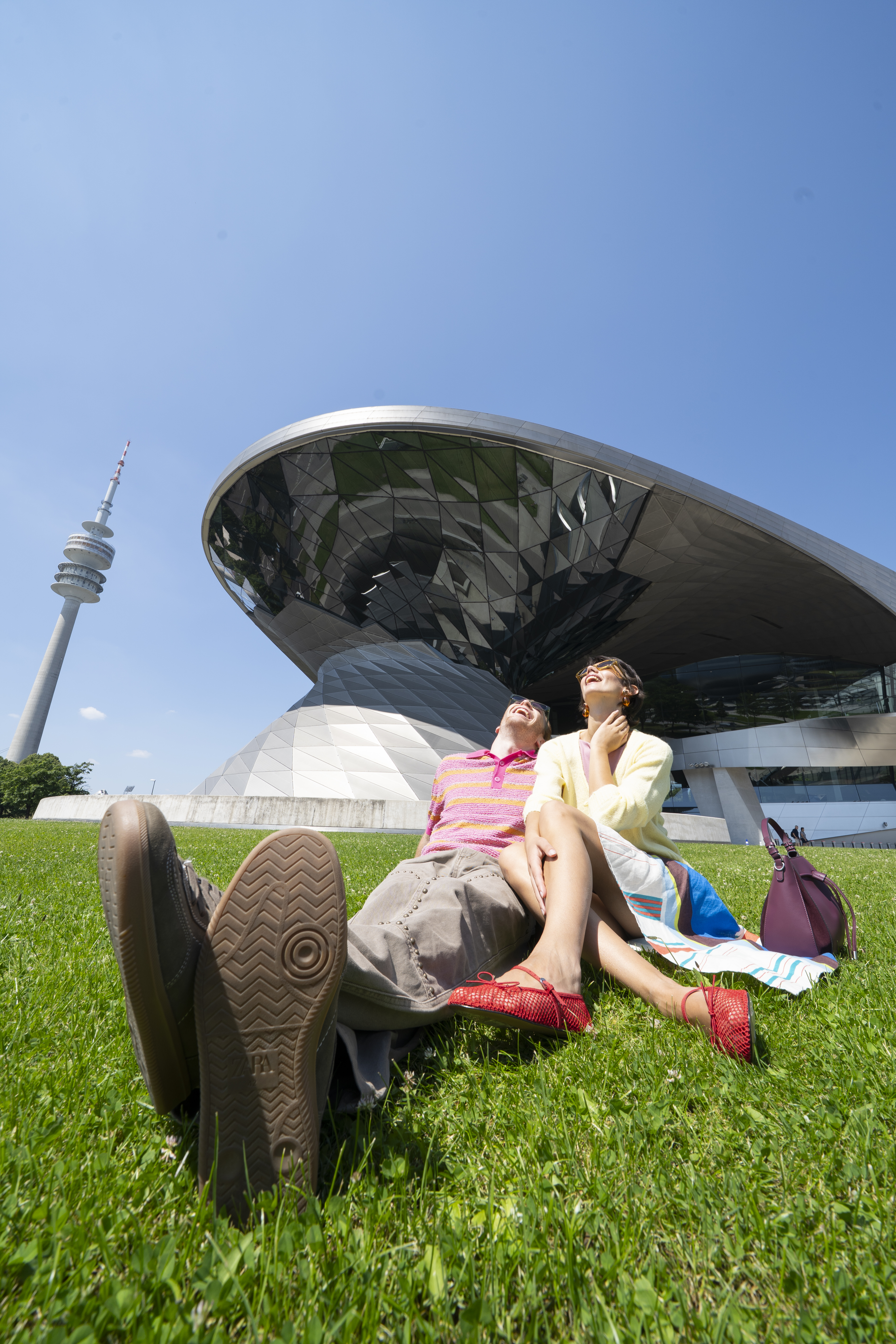 Happy couple relaxing on grass with BMW Welt and Olympiaturm in Munich.