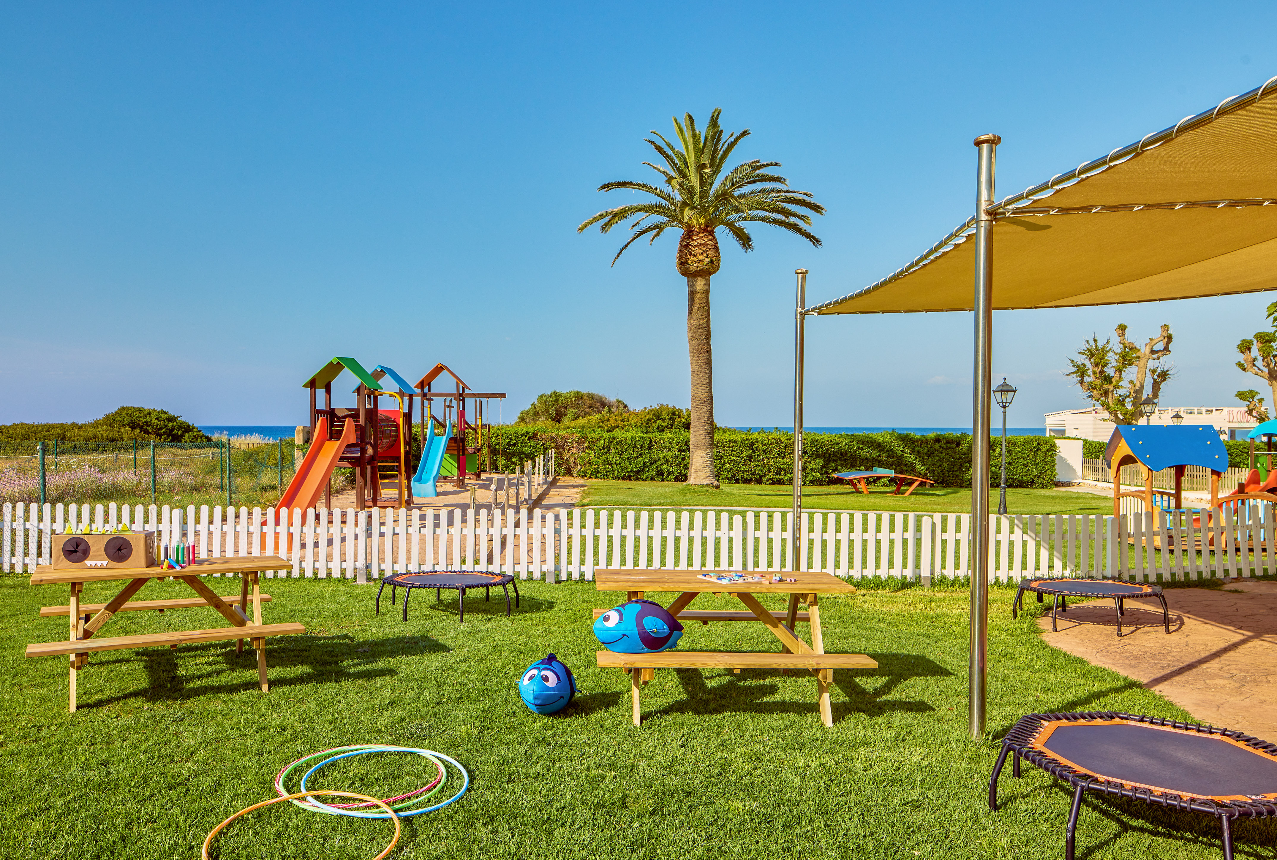 Sunny outdoor playground with slides, trampolines, picnic tables, palm tree, ocean.