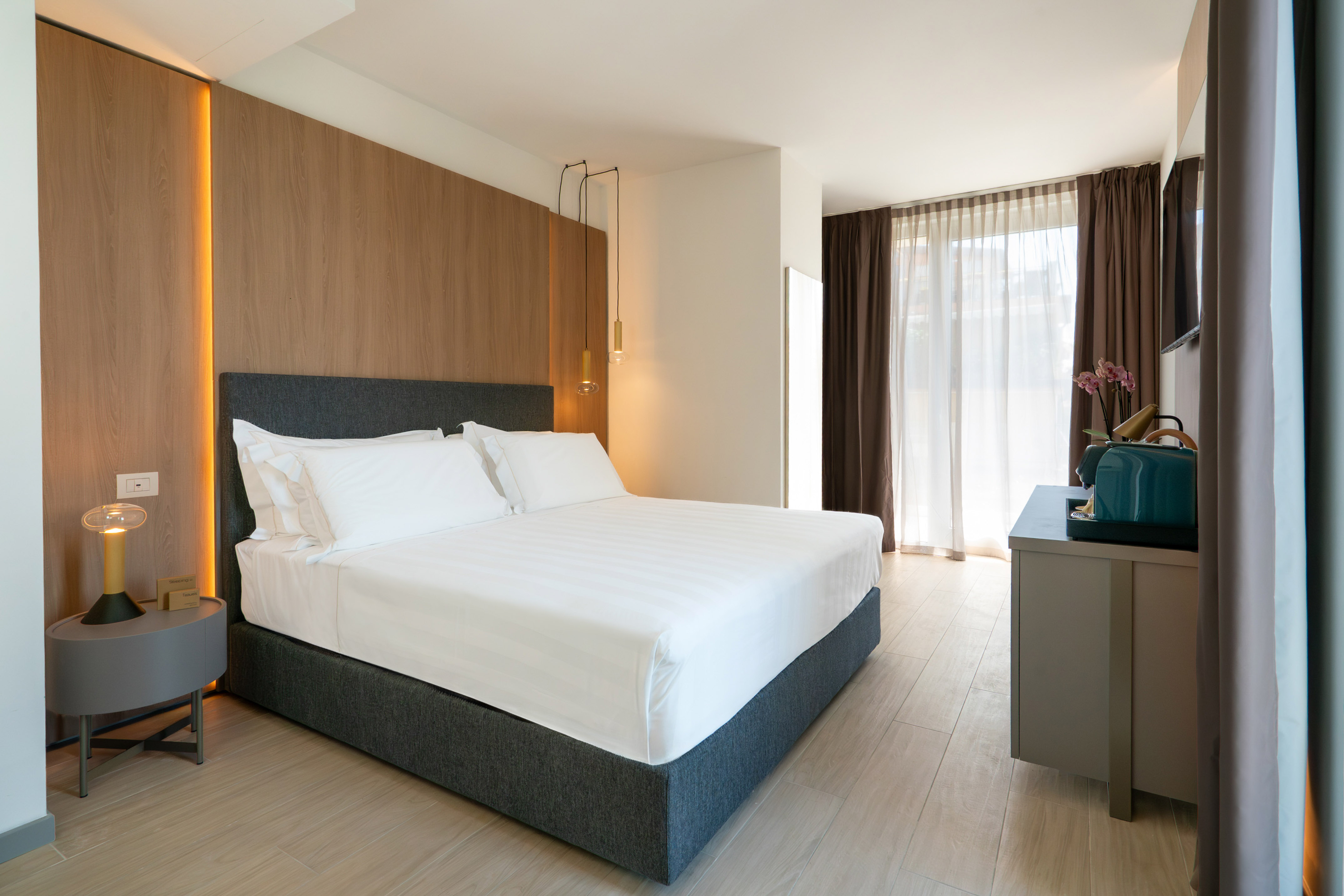 Modern hotel room with white bed, wooden accent wall, and bright window.