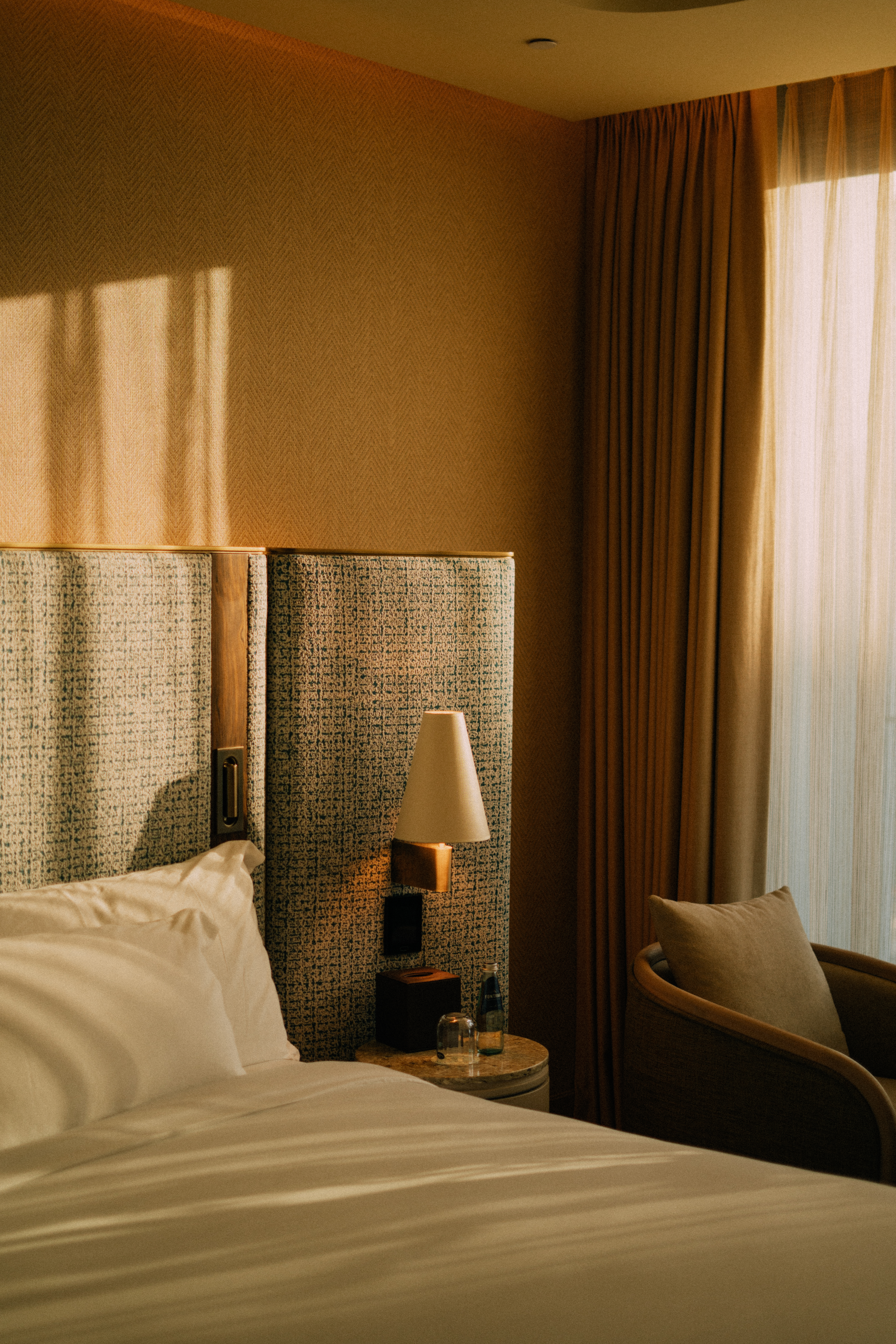 Warm hotel bedroom with bed, white linens, bedside lamp, and sunny window.