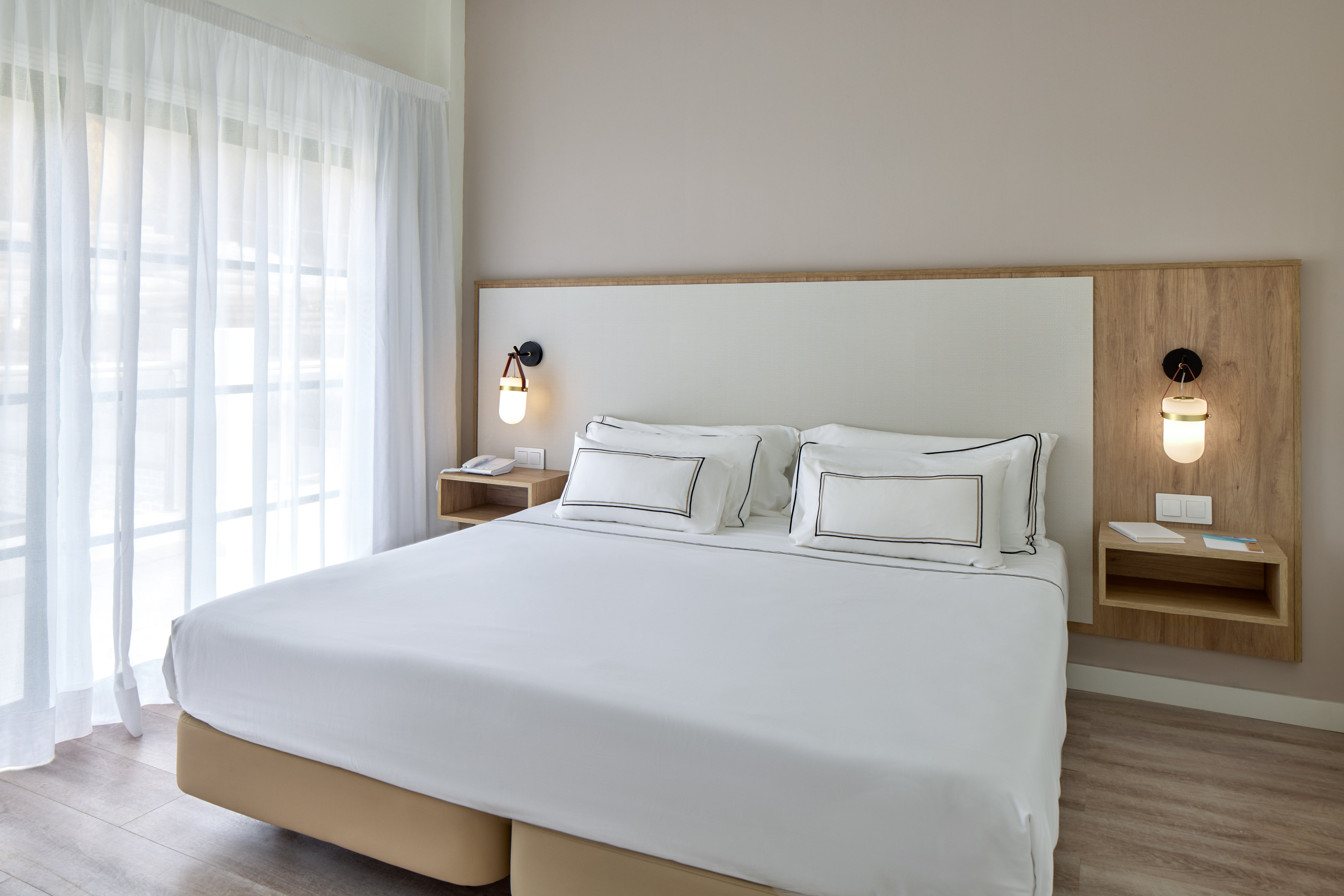 Clean hotel bedroom with white bed, wood accents, and window curtains.
