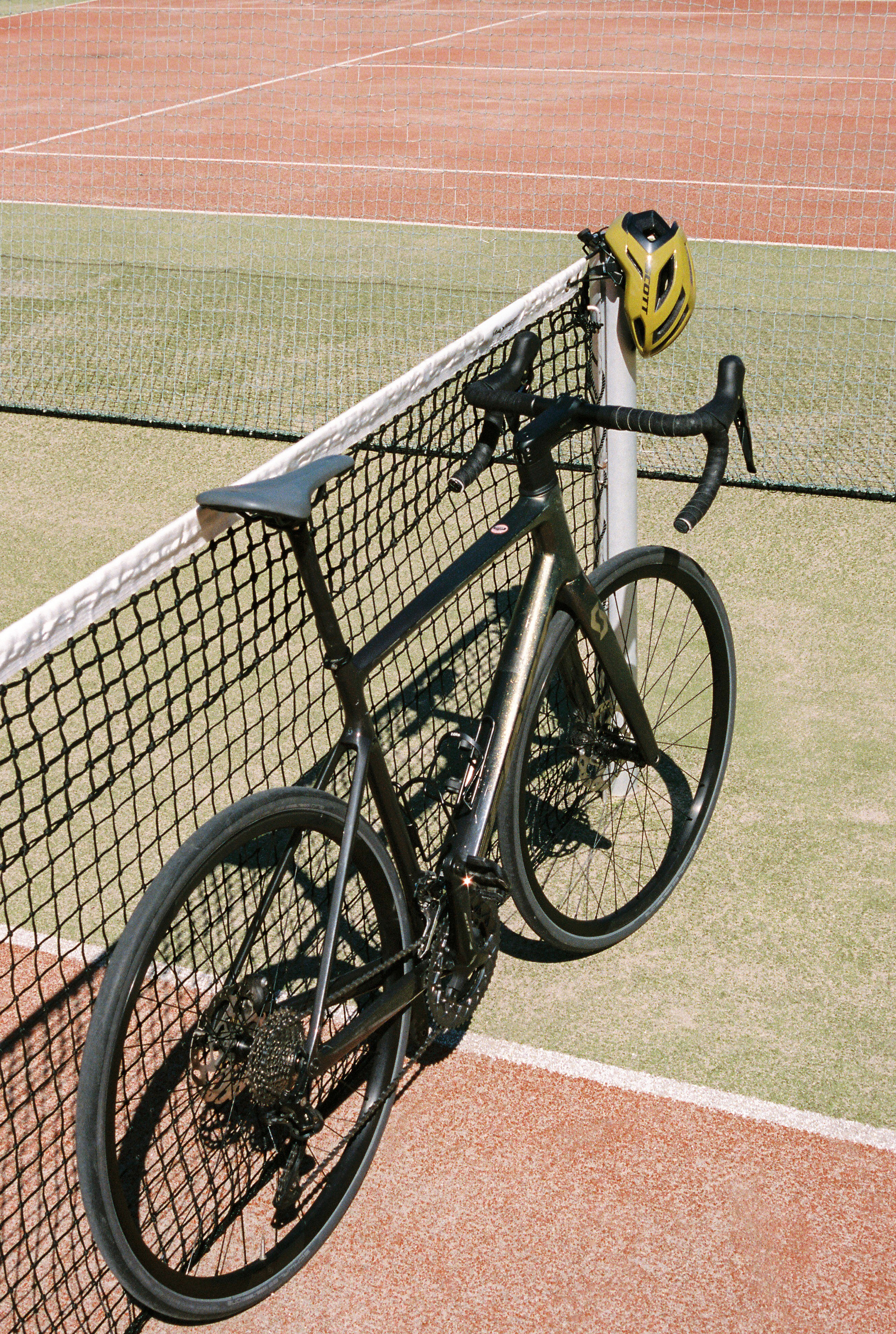 Dark Scott road bike & yellow helmet by tennis court net.