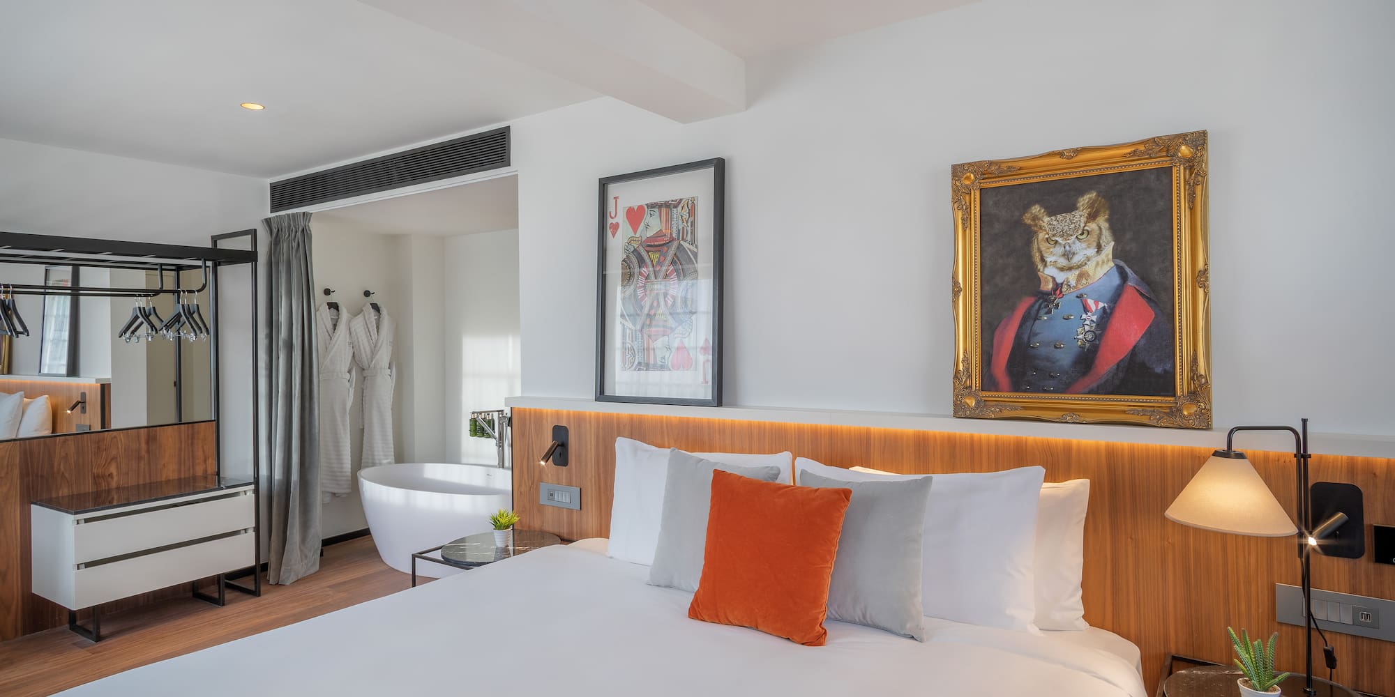 Open-plan hotel room with king bed, freestanding tub, and quirky wall art.