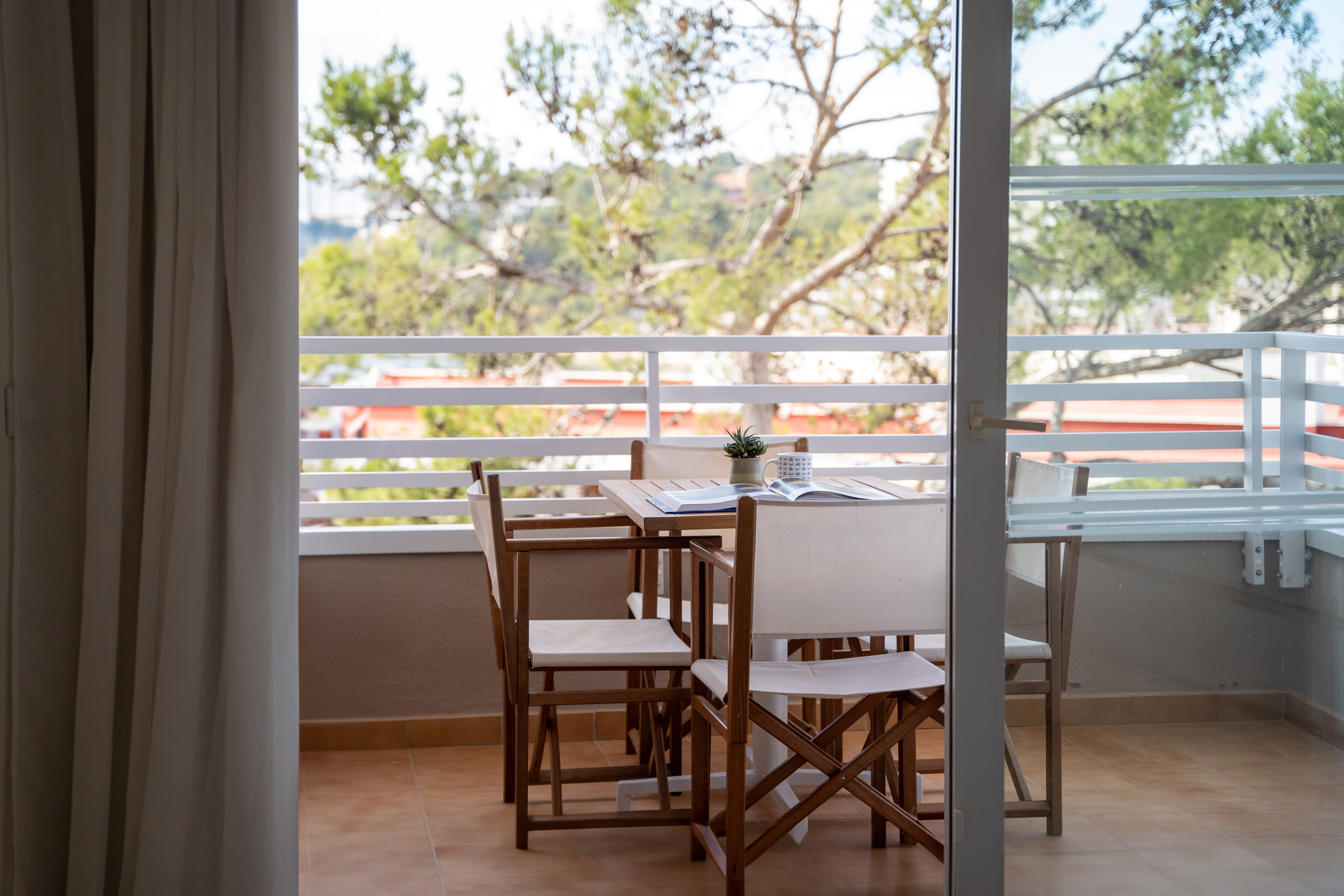 Outdoor balcony with wooden table, chairs, plant, and scenic tree view.