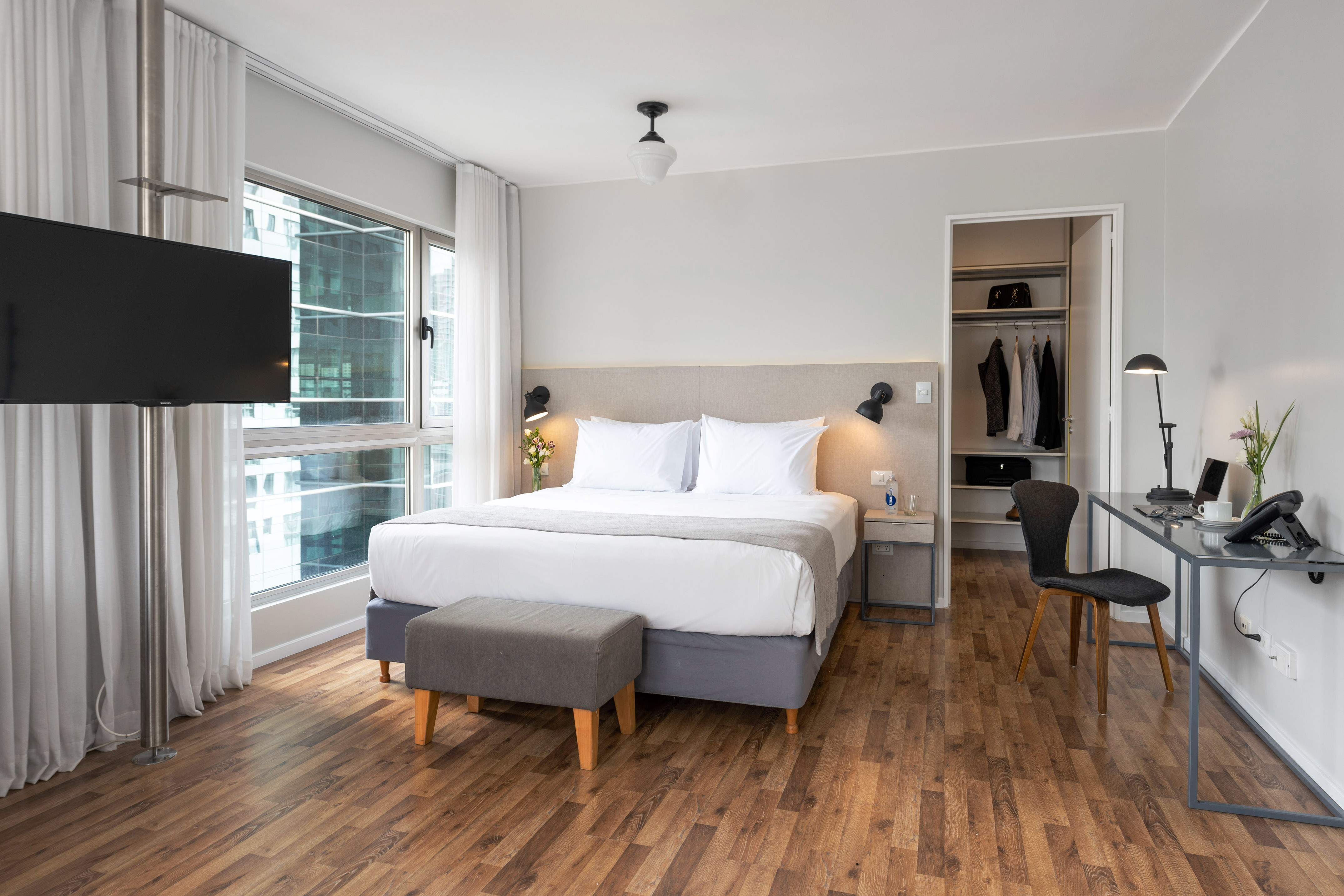 Modern hotel suite with large bed, city view, desk, TV, and open wardrobe.