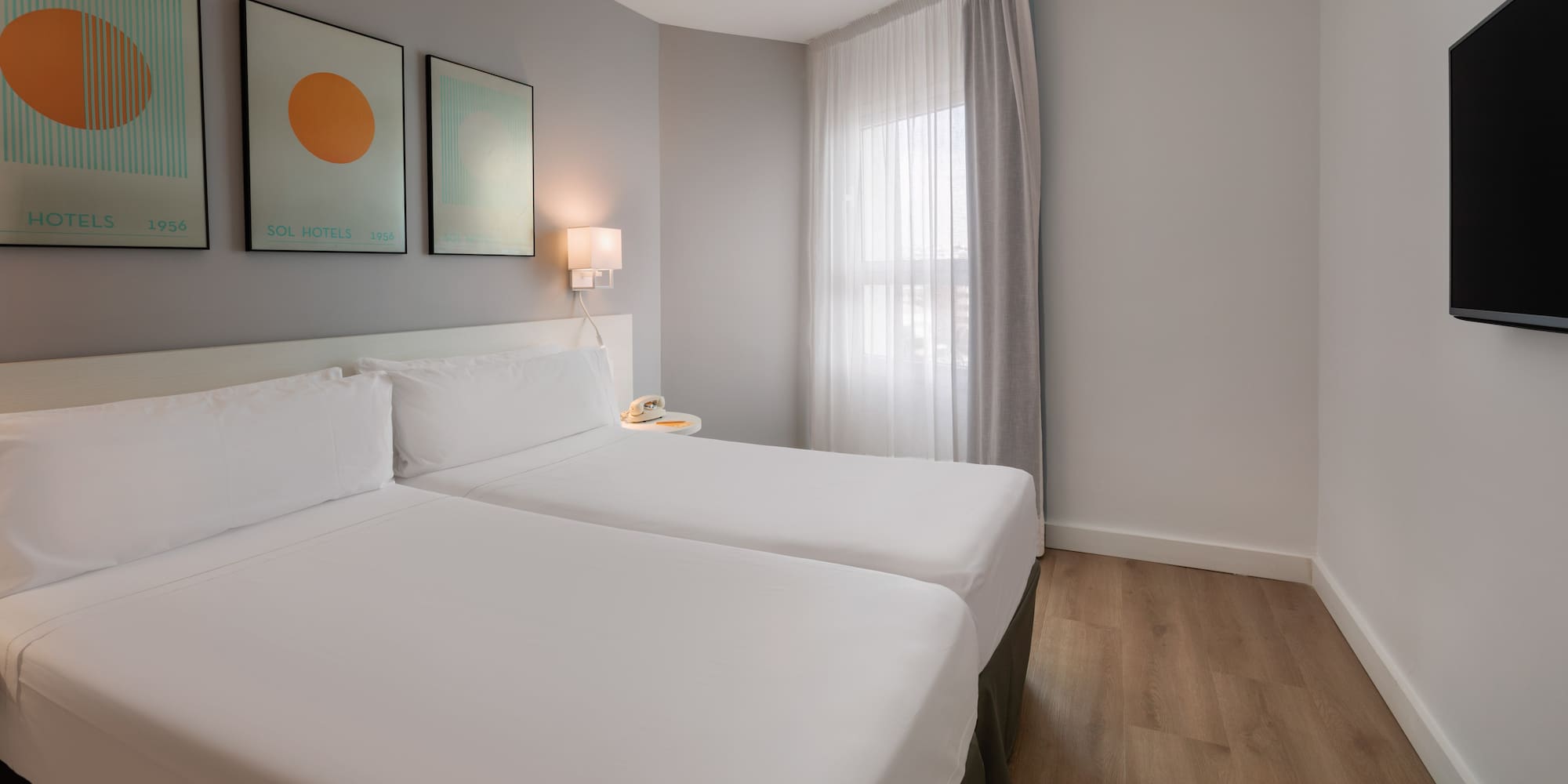 Modern hotel twin room with white beds, wood floor, abstract art, and window.