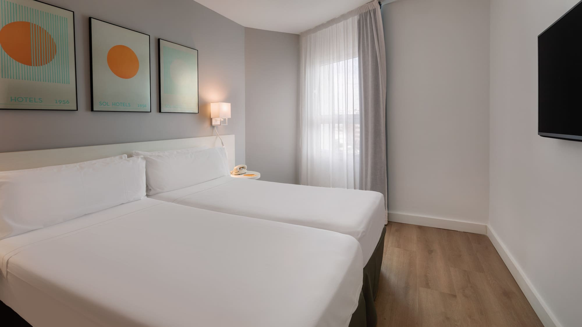 Modern hotel twin room with white beds, wood floor, abstract art, and window.