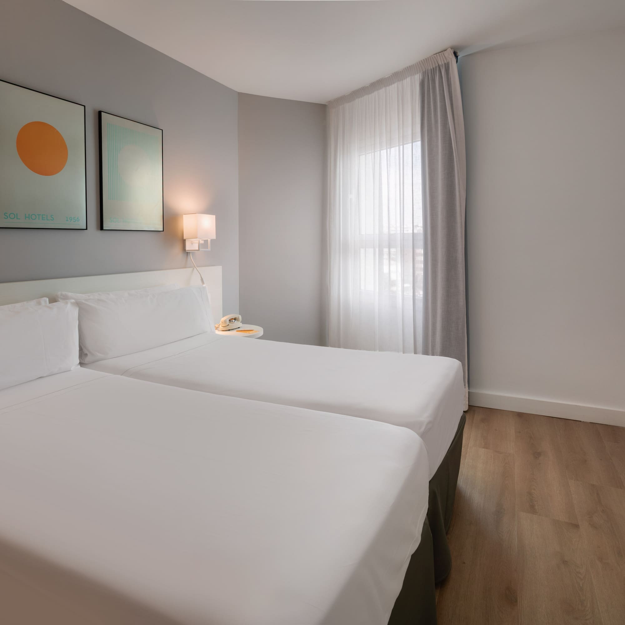 Modern hotel twin room with white beds, wood floor, abstract art, and window.