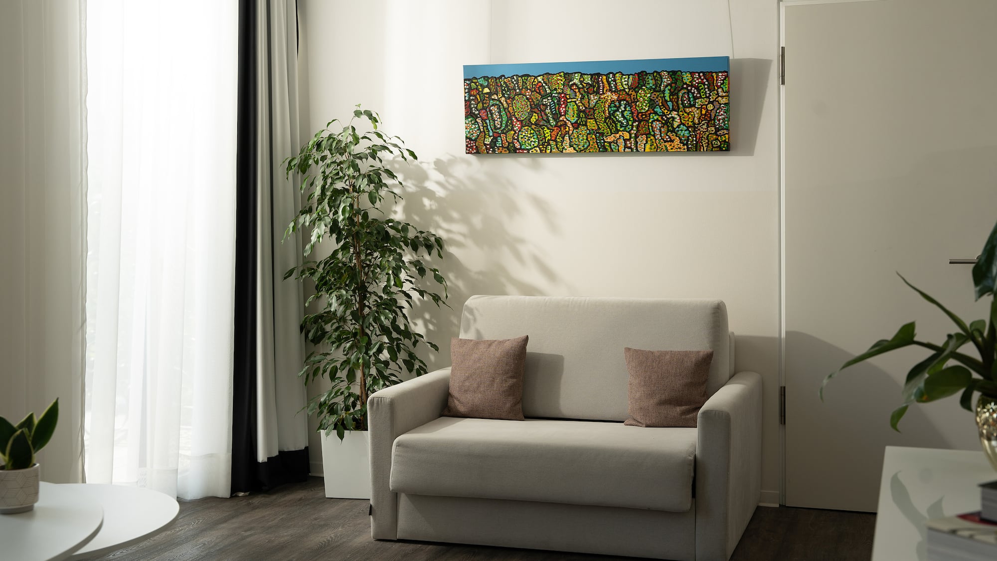 Modern living room with grey sofa, colorful abstract painting, and tall ficus plant.