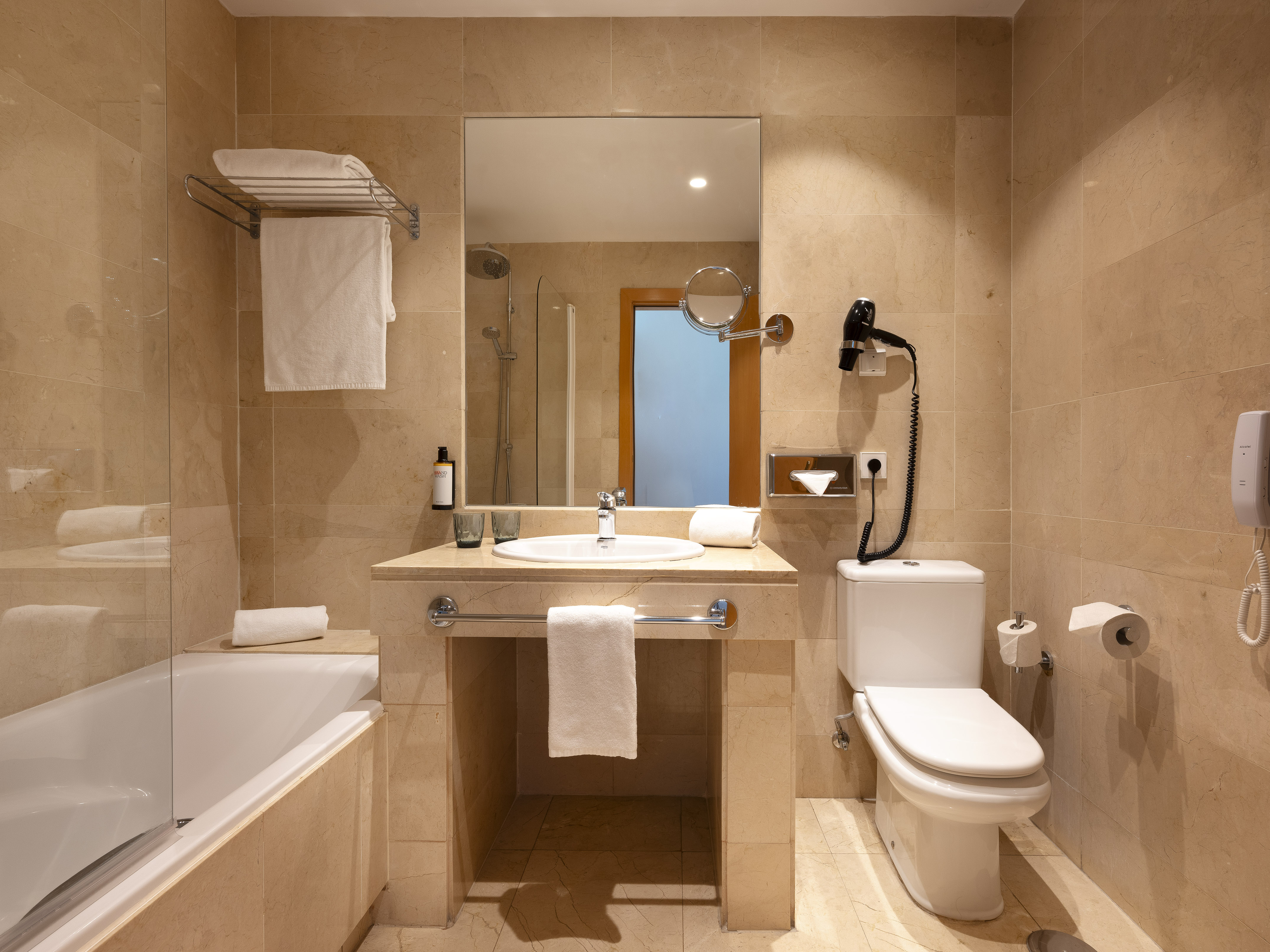 Modern hotel bathroom with beige tiled walls, bathtub, sink, toilet, and white towels.