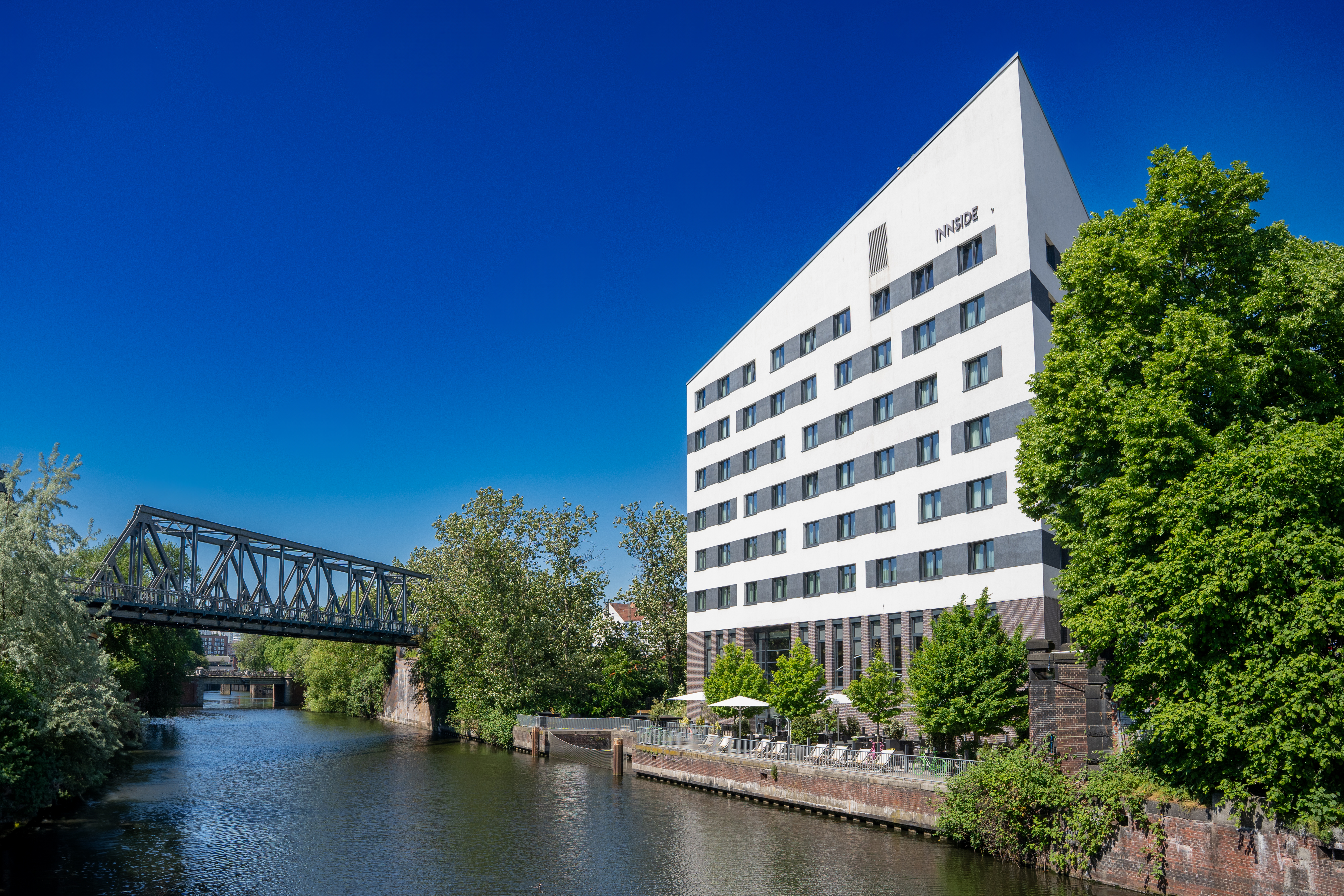 Innside hotel by river, modern architecture, metal bridge, green trees.