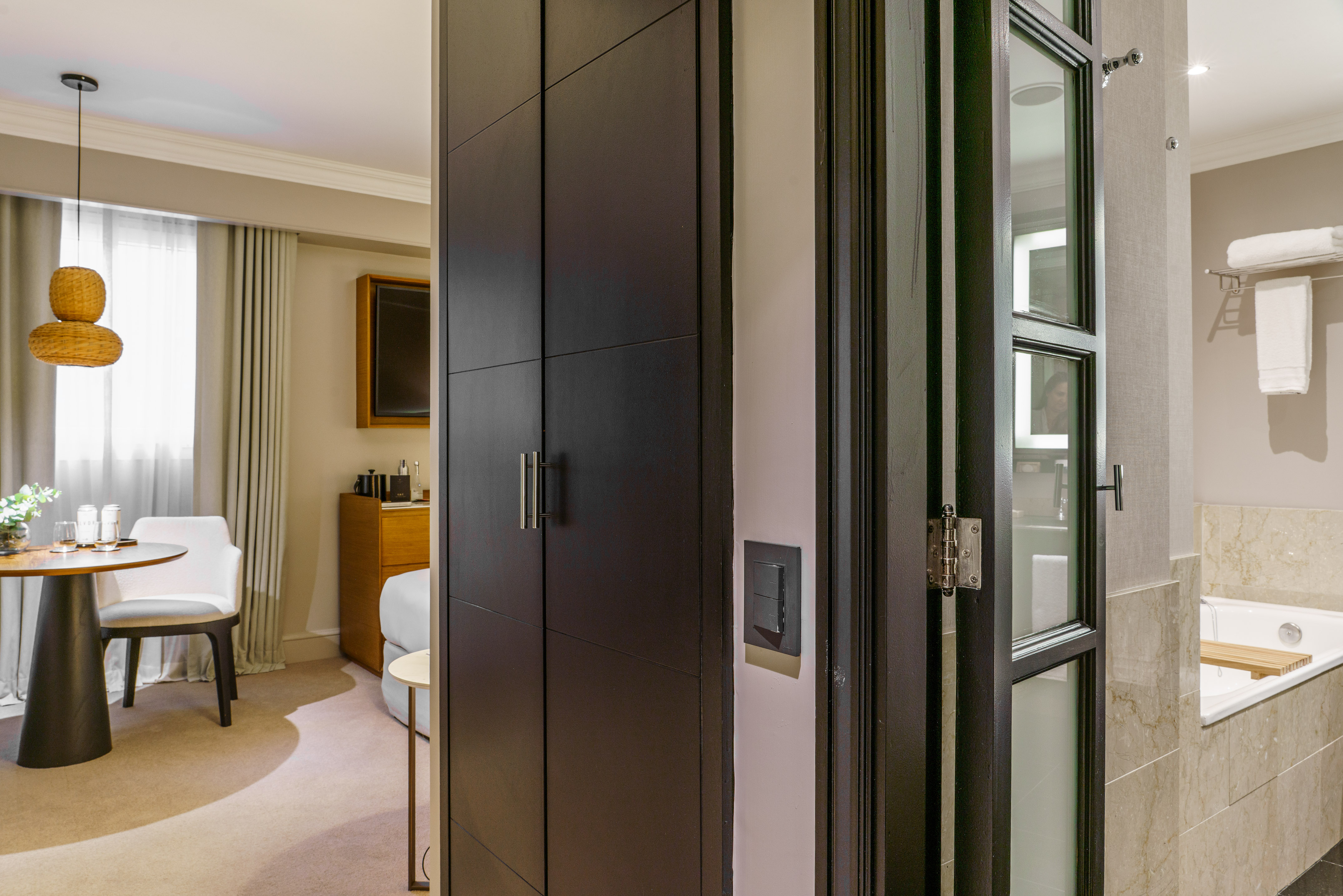 Hotel suite interior with dark wardrobe, dining area, and bathroom with bathtub.