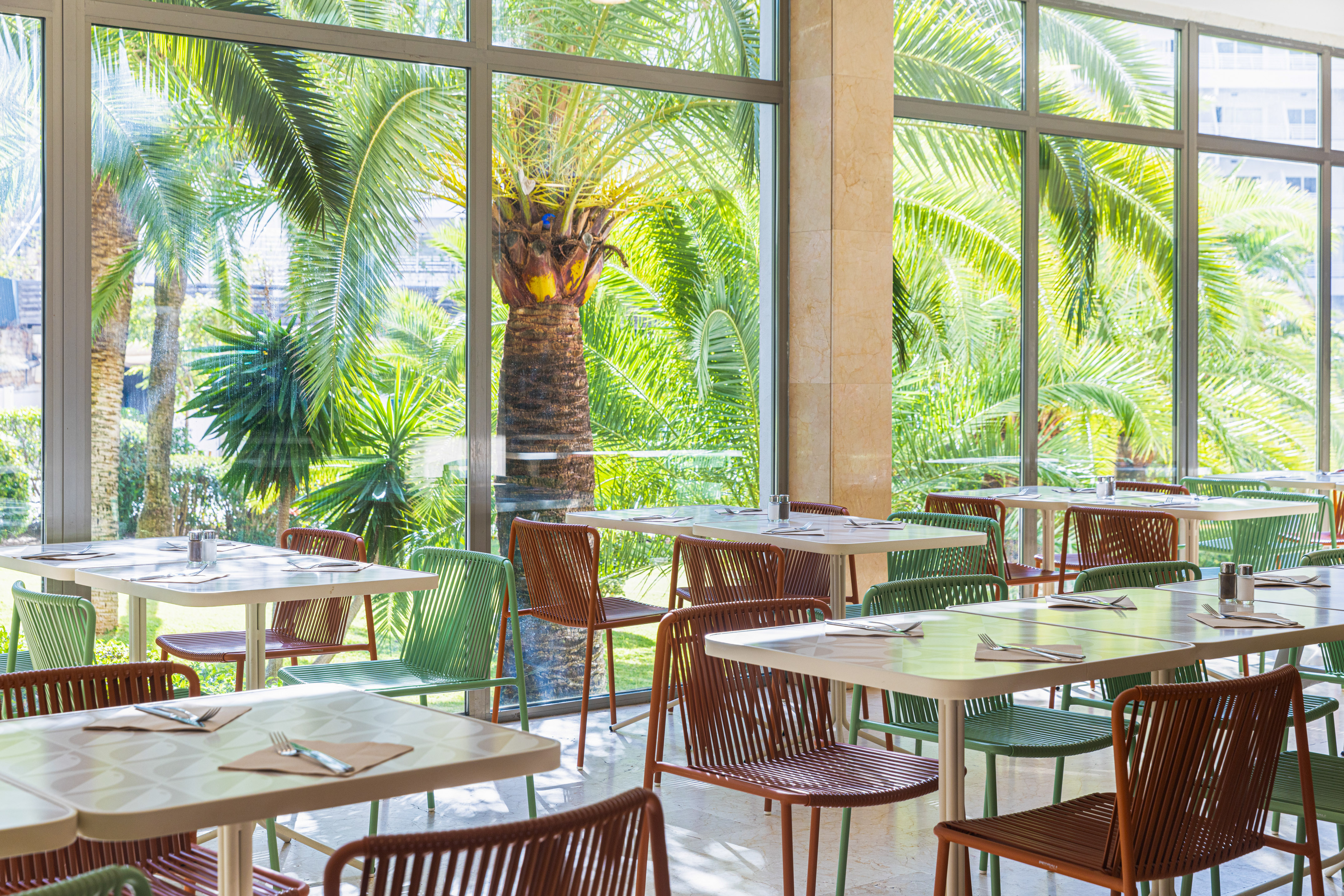Hotel restaurant dining room with bright windows overlooking lush palm trees.