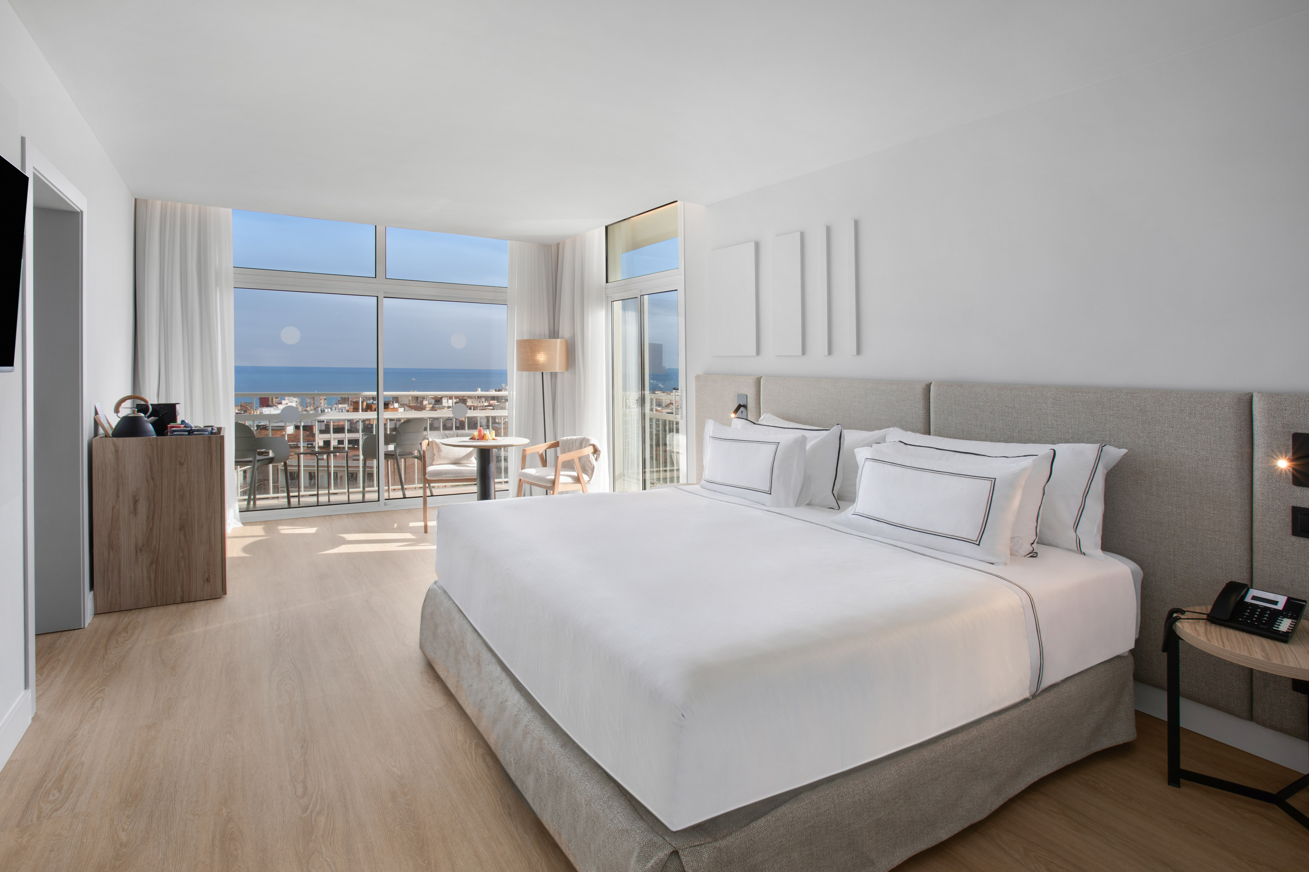 Bright modern hotel bedroom with sea view, balcony, and white bed.