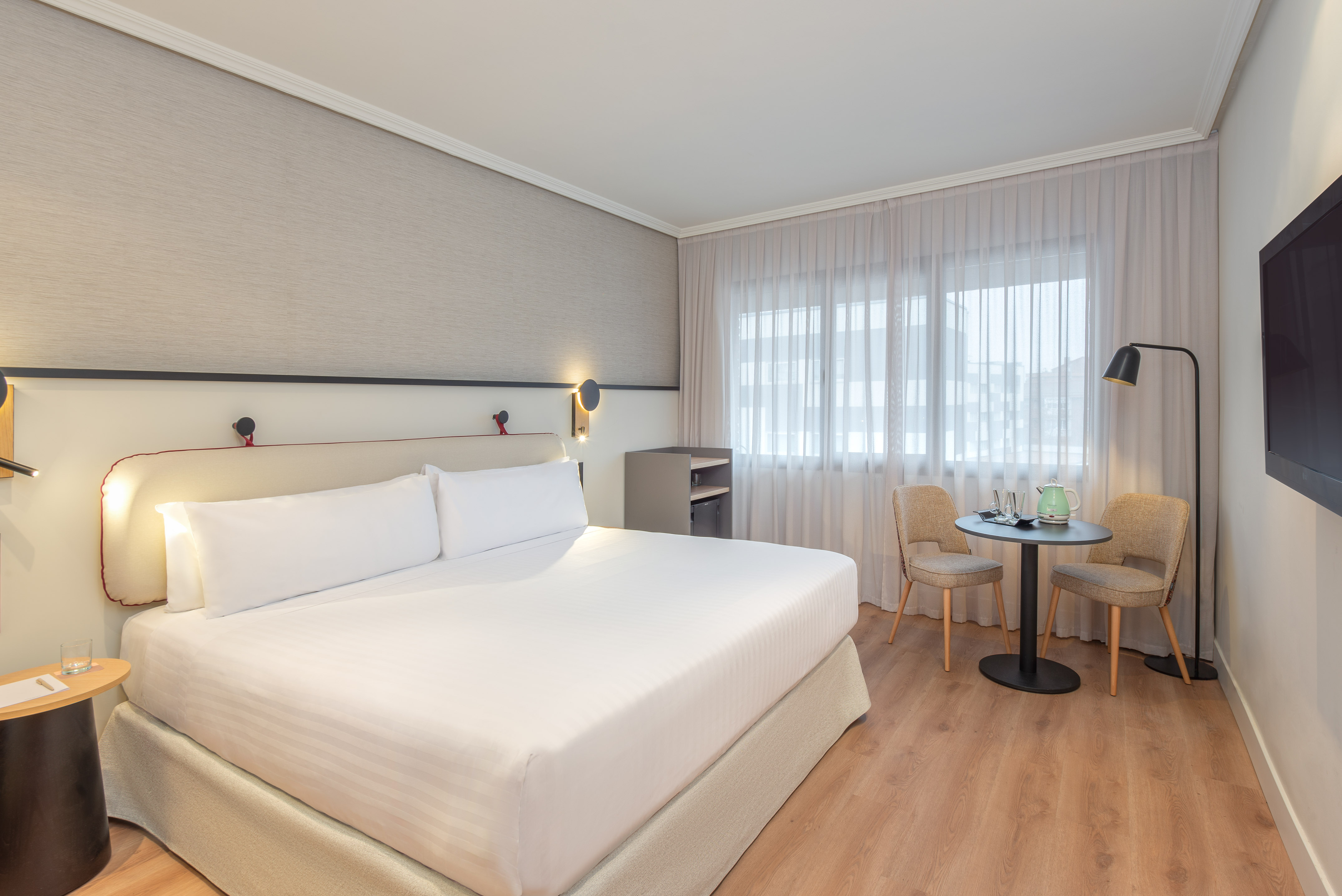 Modern hotel room with white bed, seating area, large window, and TV.