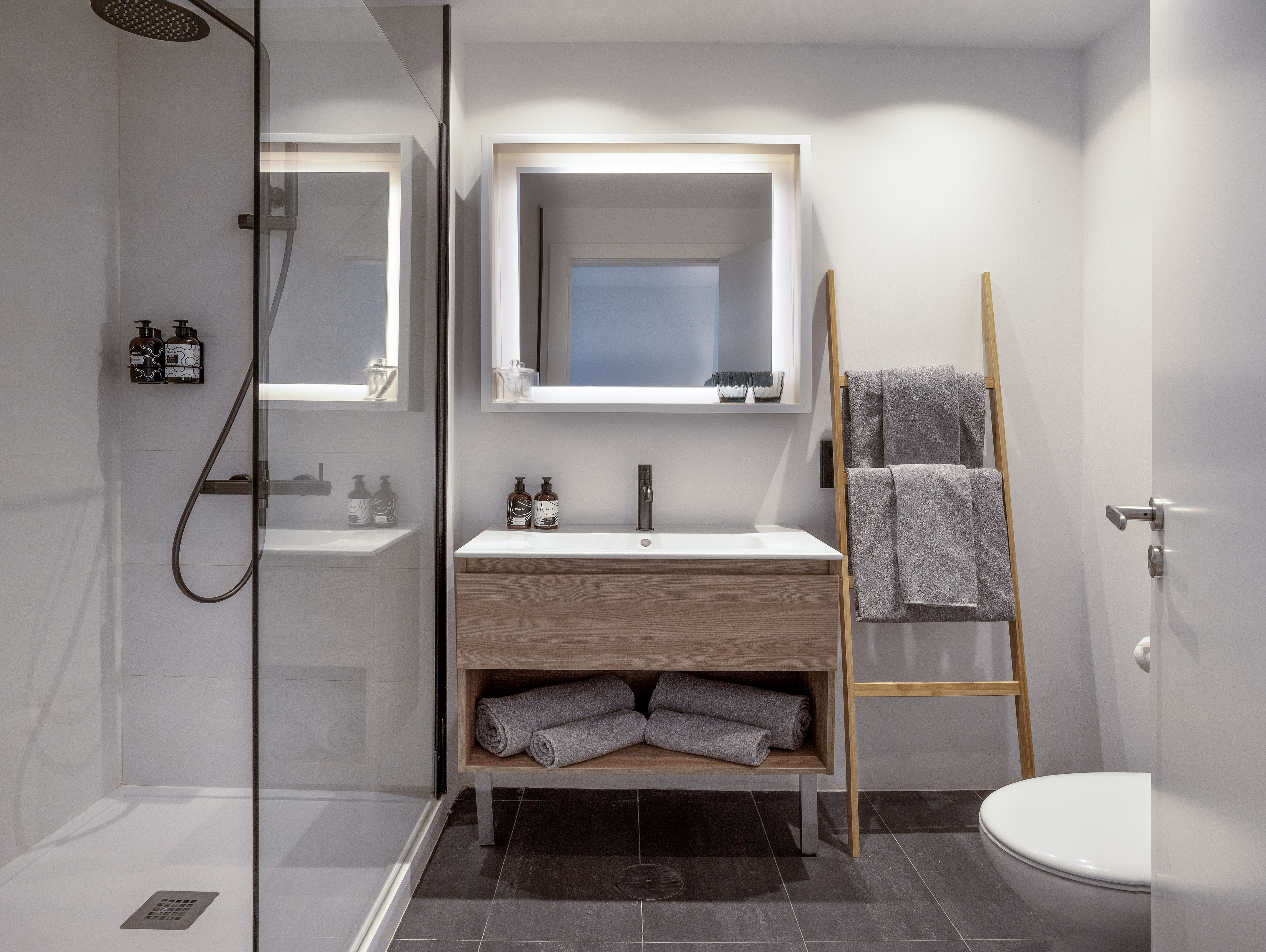 Modern bathroom with glass shower, wooden vanity, and illuminated mirror.
