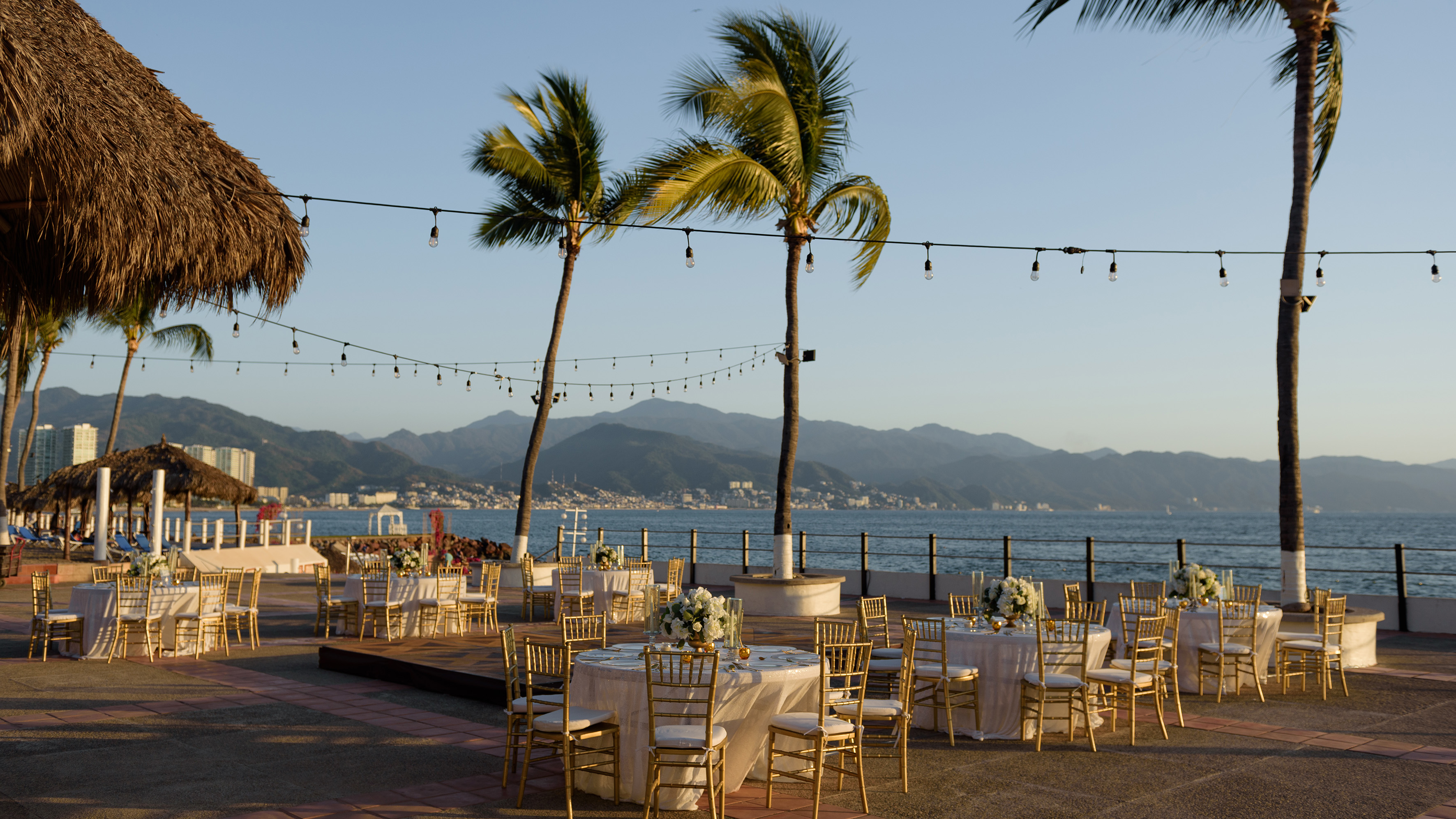 Elegant waterfront dining tables, gold chairs, palm trees, string lights.