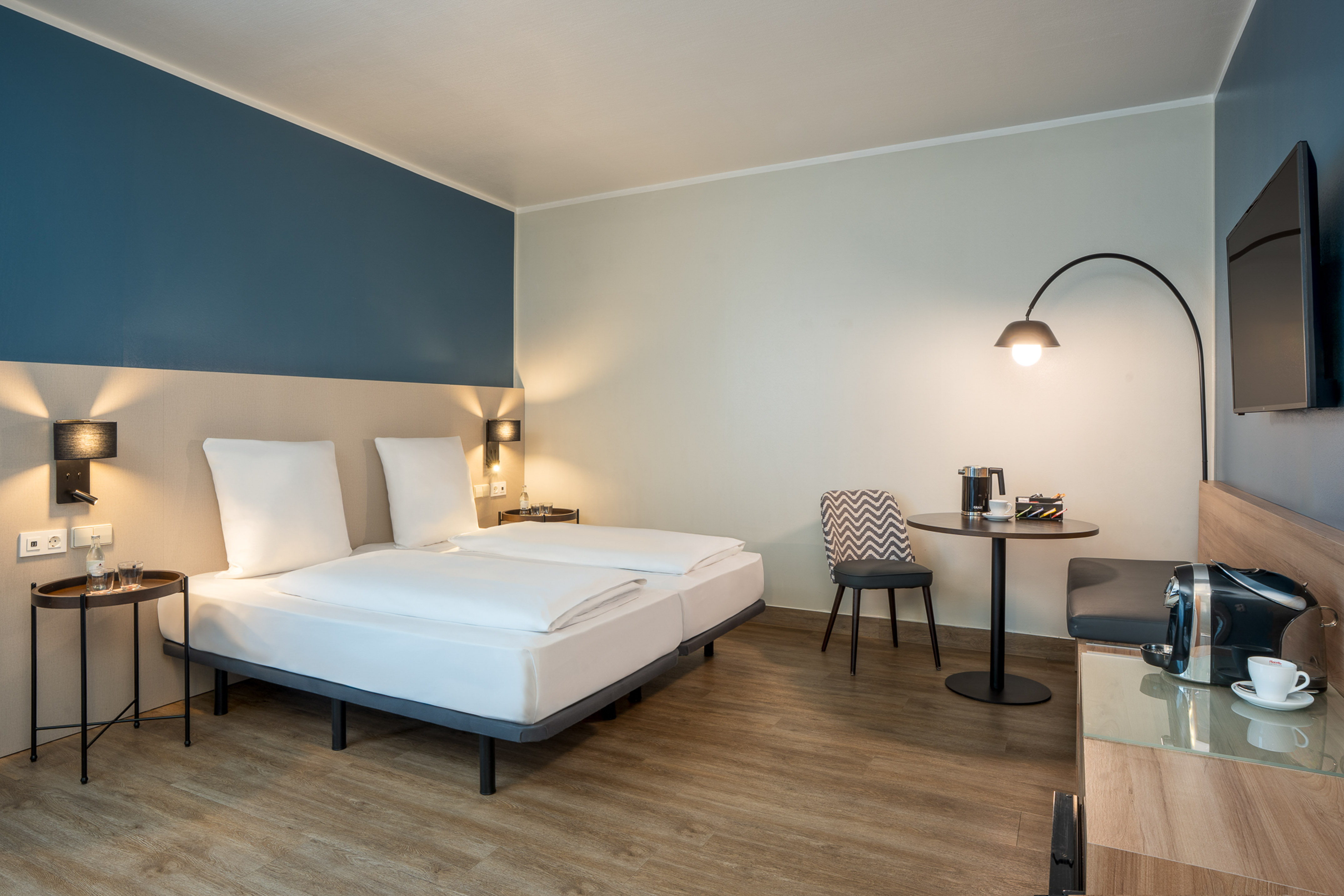 Modern hotel room with two beds, blue accent wall, coffee station, and TV.