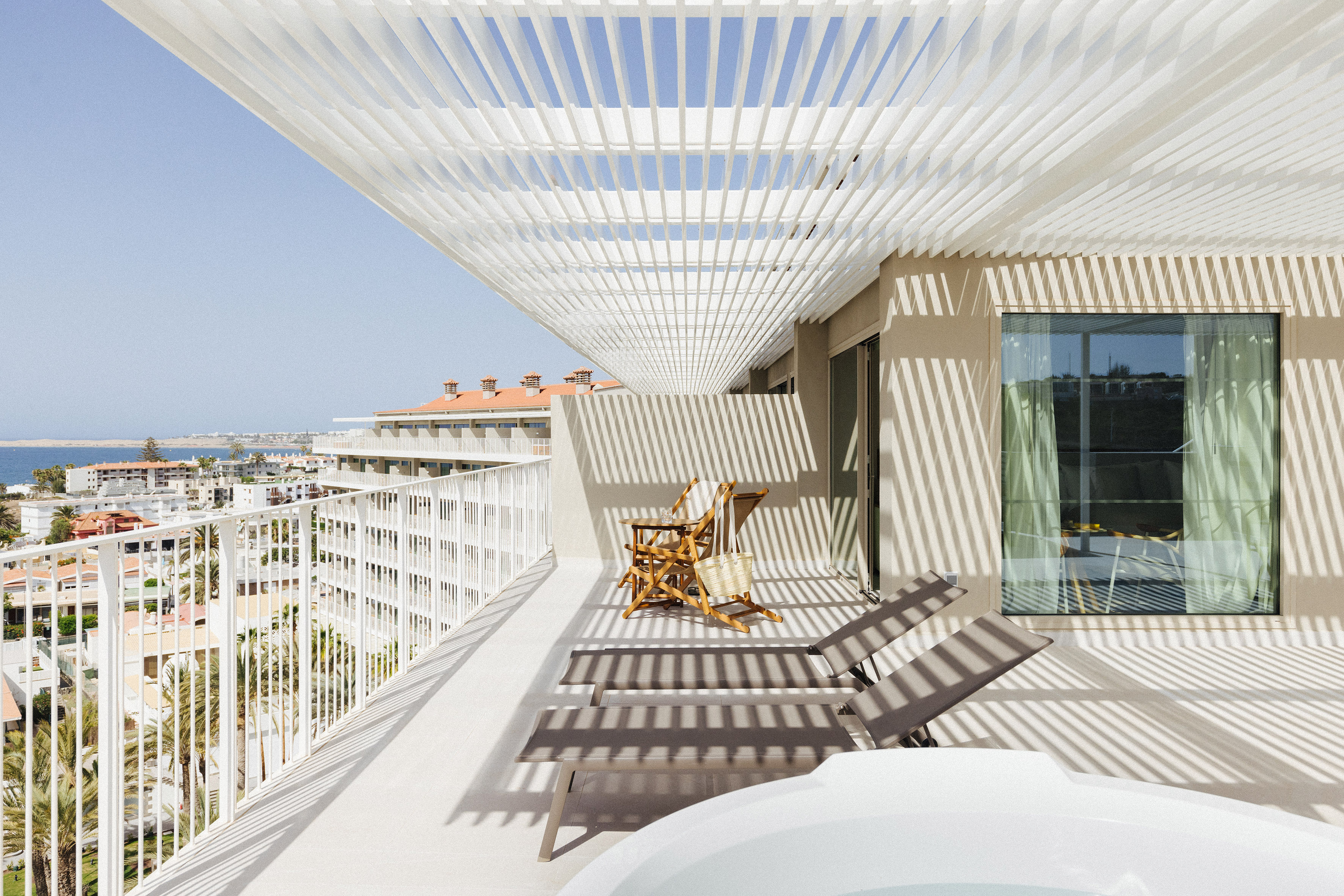 Modern hotel balcony with sea view, slatted pergola, sun loungers, hot tub.