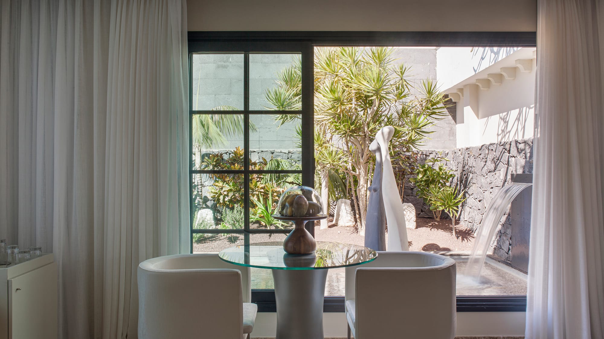 Interior moderno con mesa de cristal, sillas y vista a jardín con cascada.