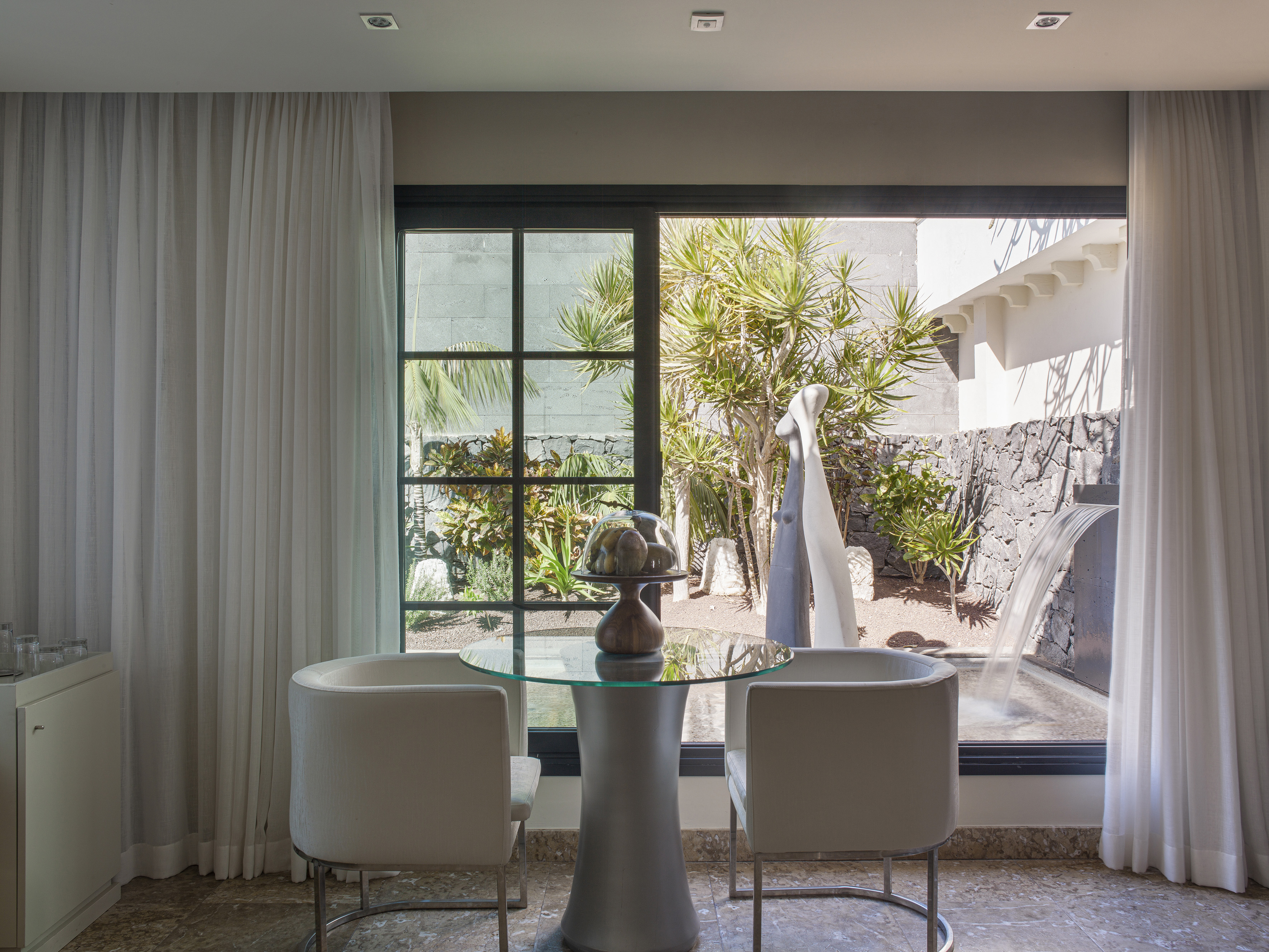 Interior moderno con mesa de cristal, sillas y vista a jardín con cascada.