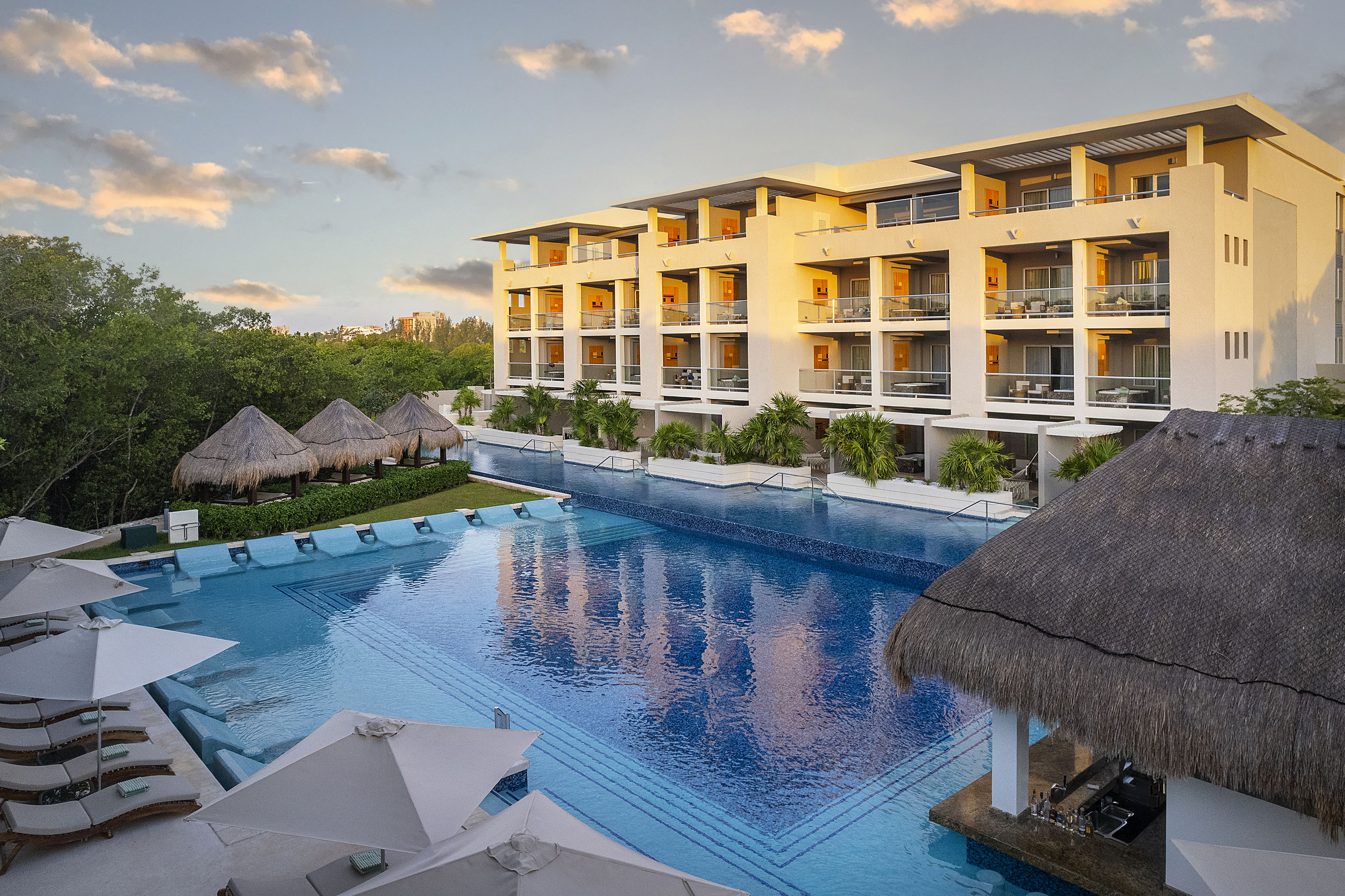 Luxury resort hotel with large pool, swim-up bar, and palapas at sunset.