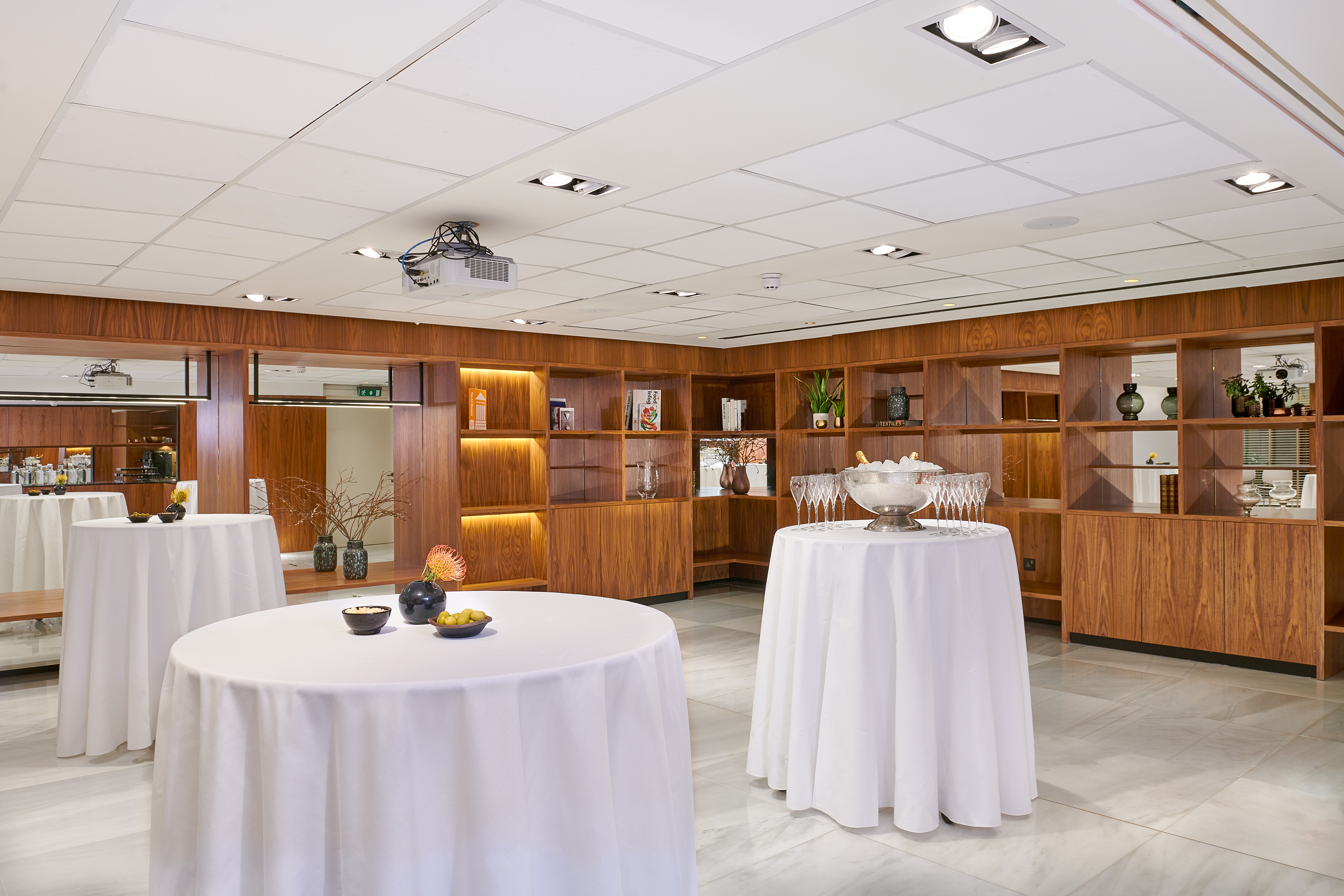 Modern reception hall with white tables, wood shelves, projector, and champagne setup.