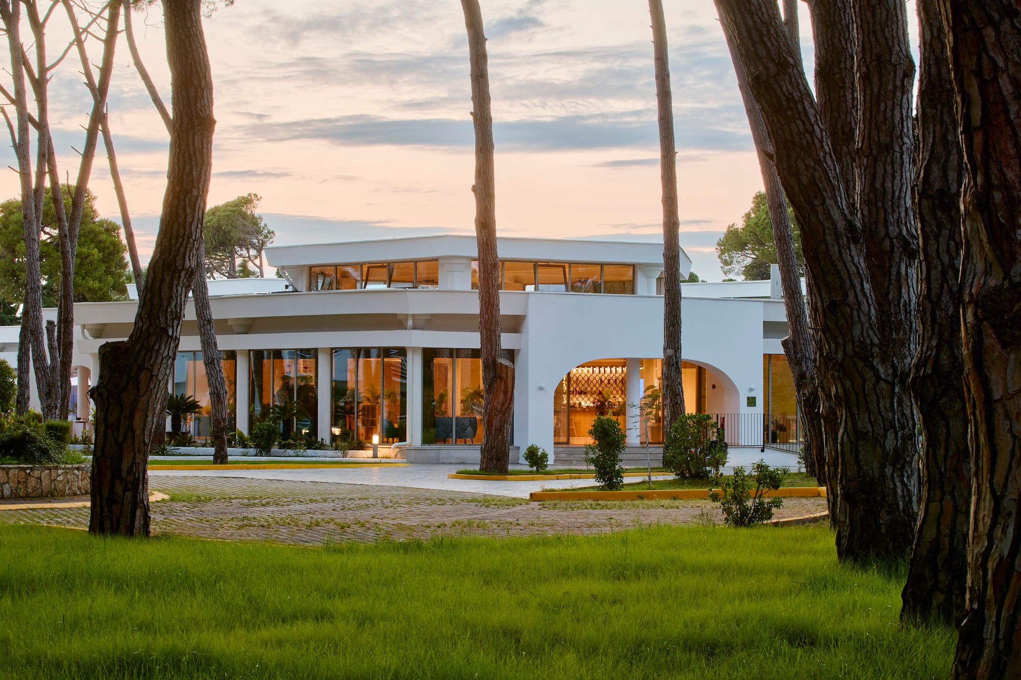 Modern white resort building at sunset, surrounded by tall pine trees.