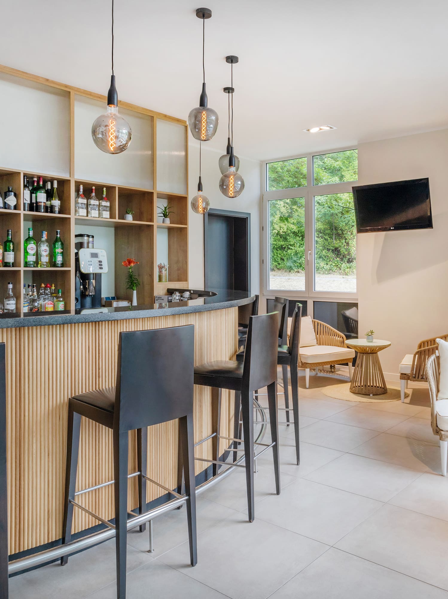 Modern hotel bar lounge with wooden counter, bar stools, and bright seating area.