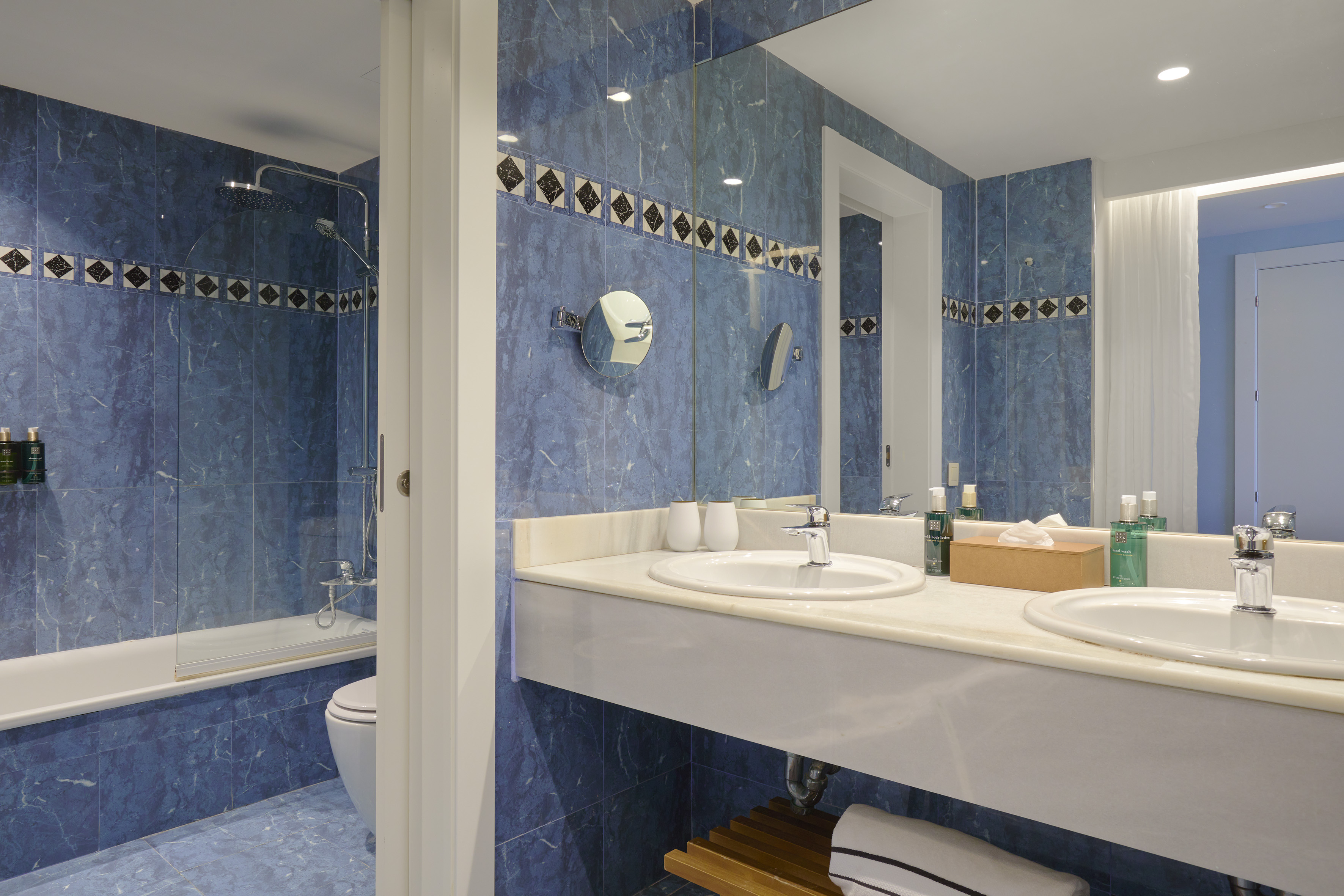 Blue marble tiled bathroom with double vanity, mirror, shower-tub, and amenities.