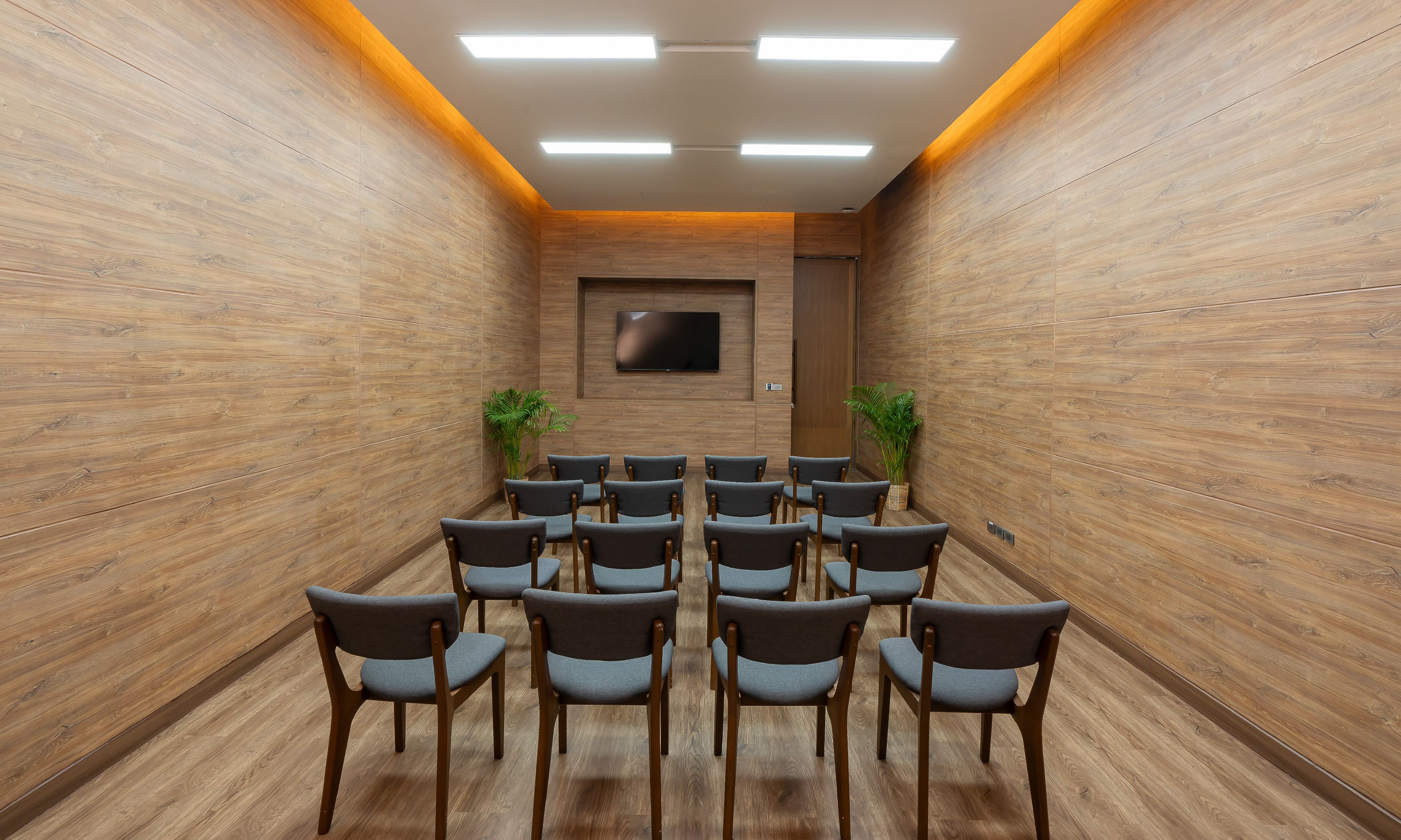 Modern conference room with wood walls, grey chairs, TV, and recessed lighting.
