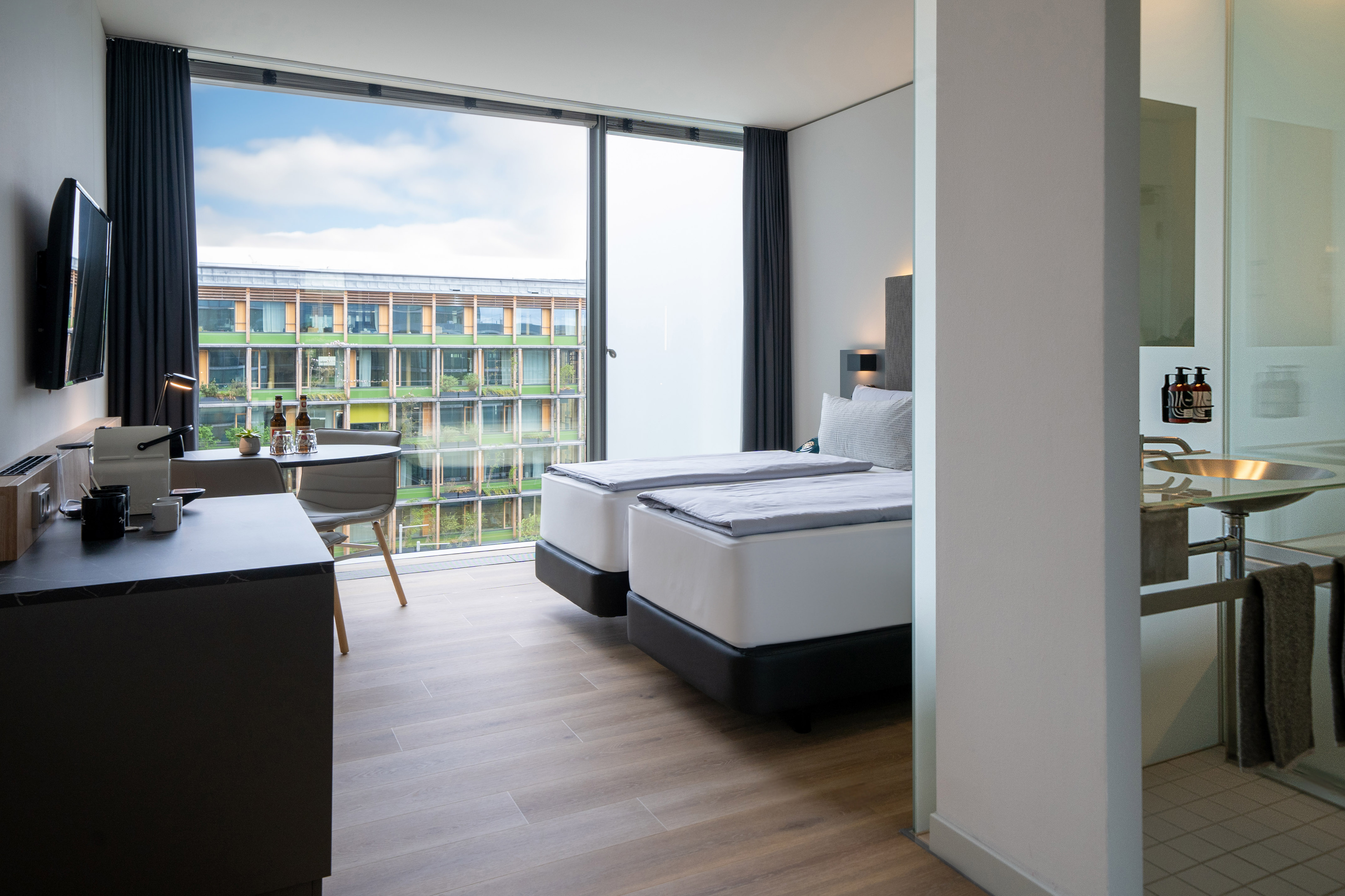 Modern hotel room with twin beds, large window, city view, and desk area.