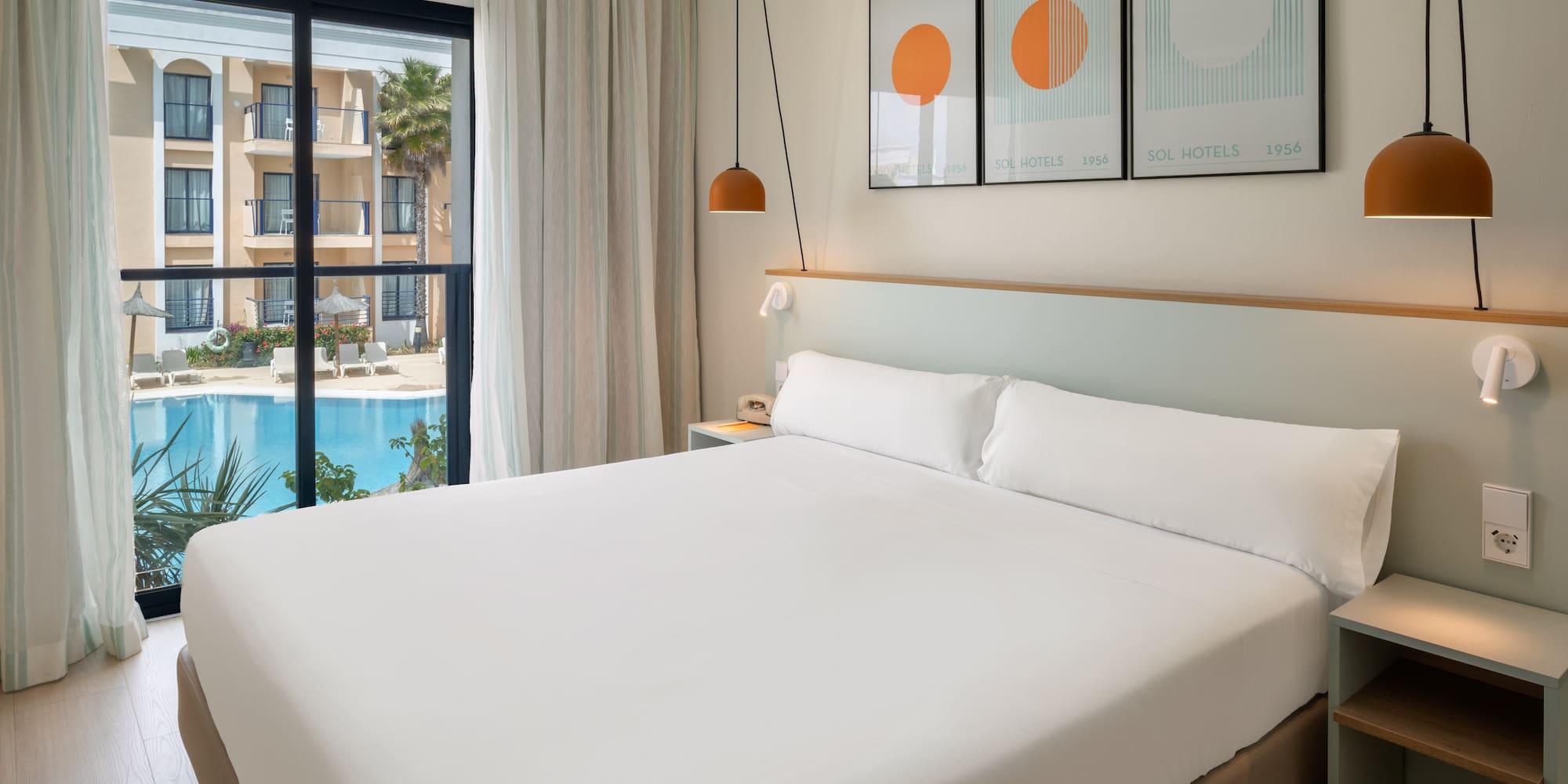 Bright hotel room with large bed, pool view, and balcony overlooking resort.