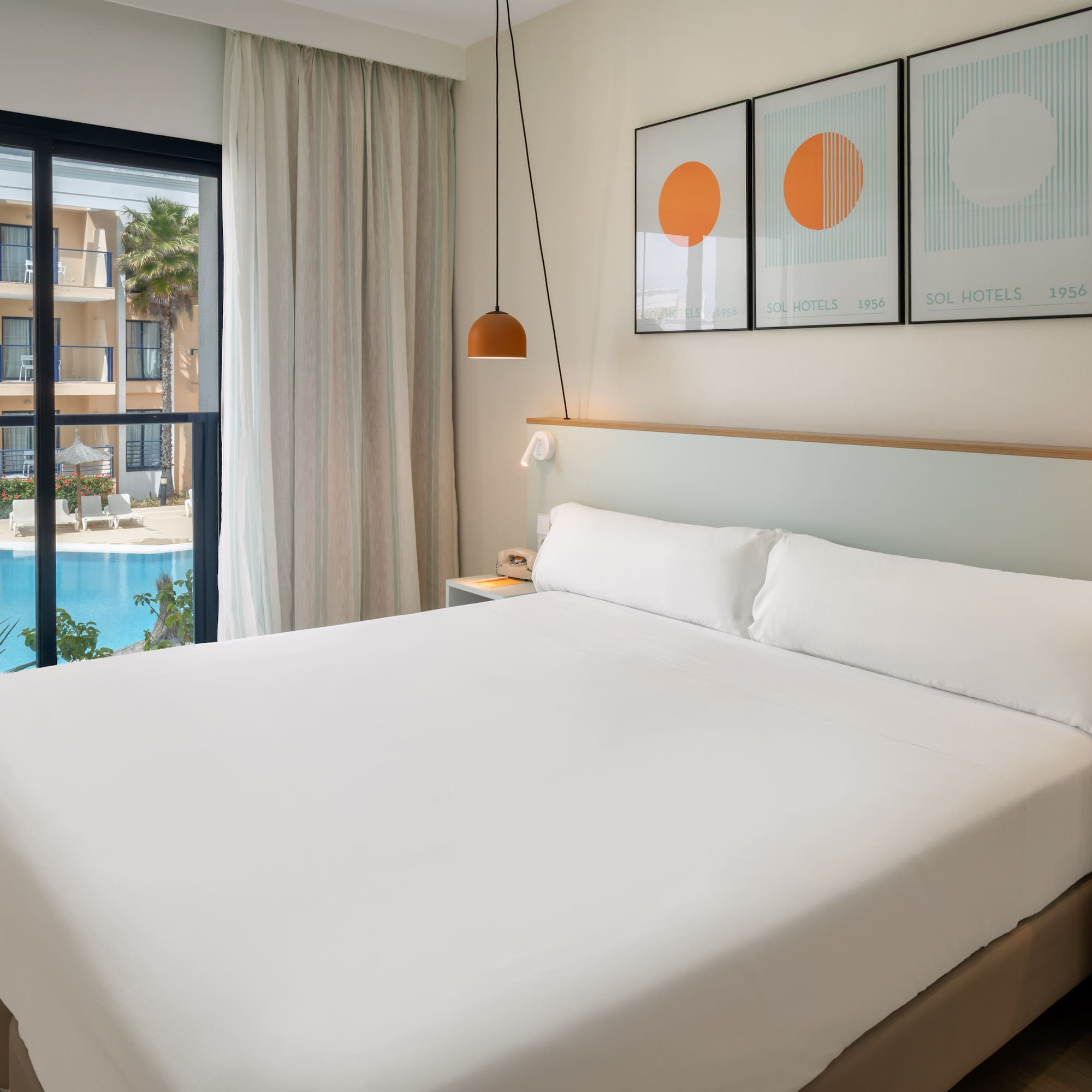 Bright hotel room with large bed, pool view, and balcony overlooking resort.