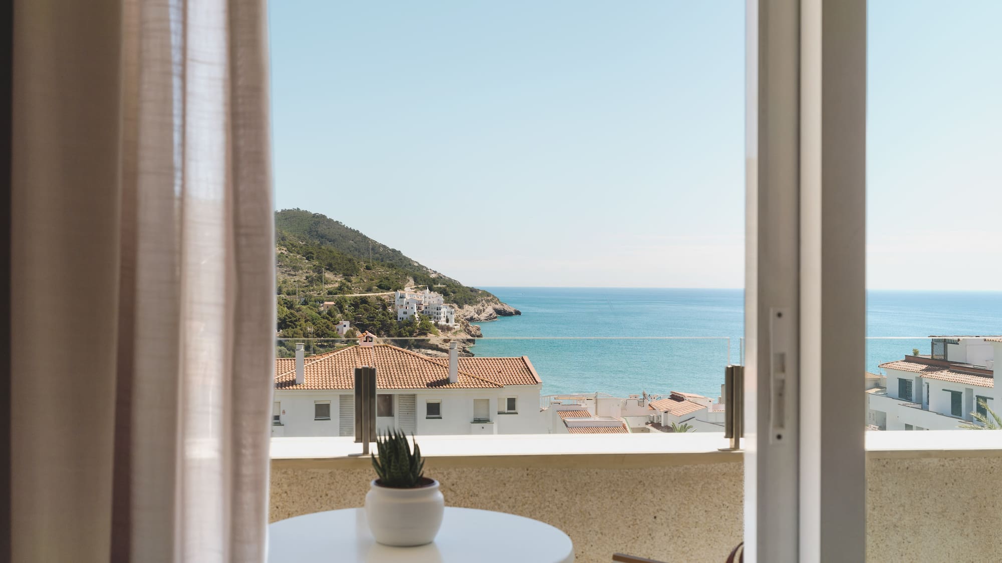 Balcony sea view: coastal village, blue water, green hills under clear sky.