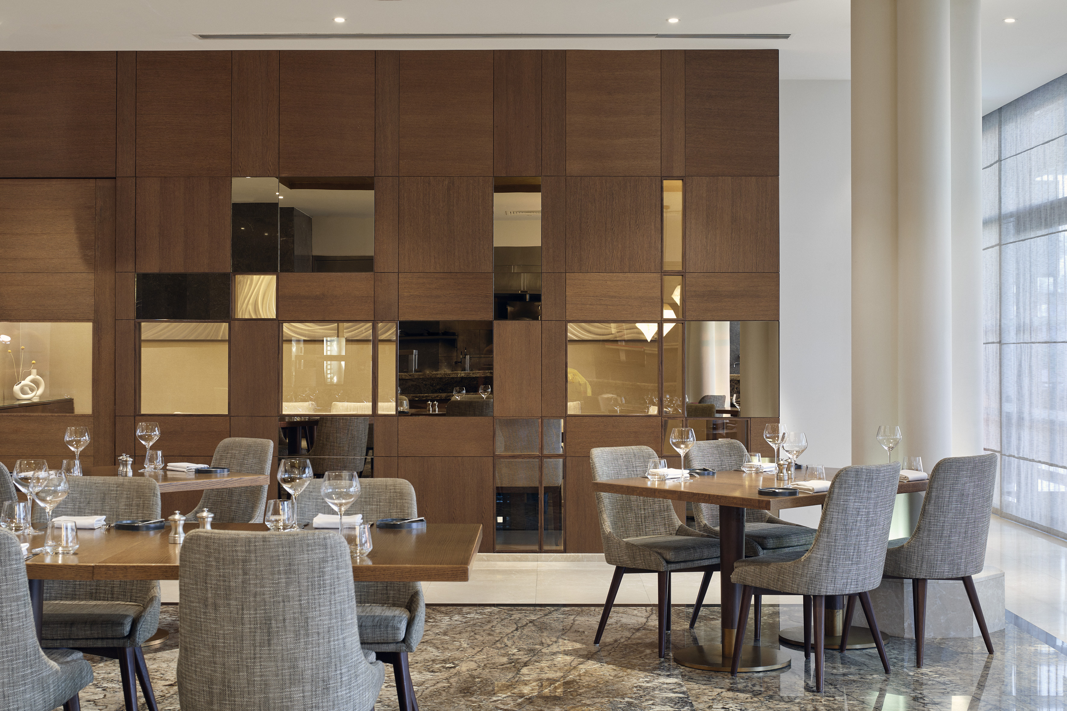 Restaurant interior with wooden tables, grey chairs, and reflective wall panels.