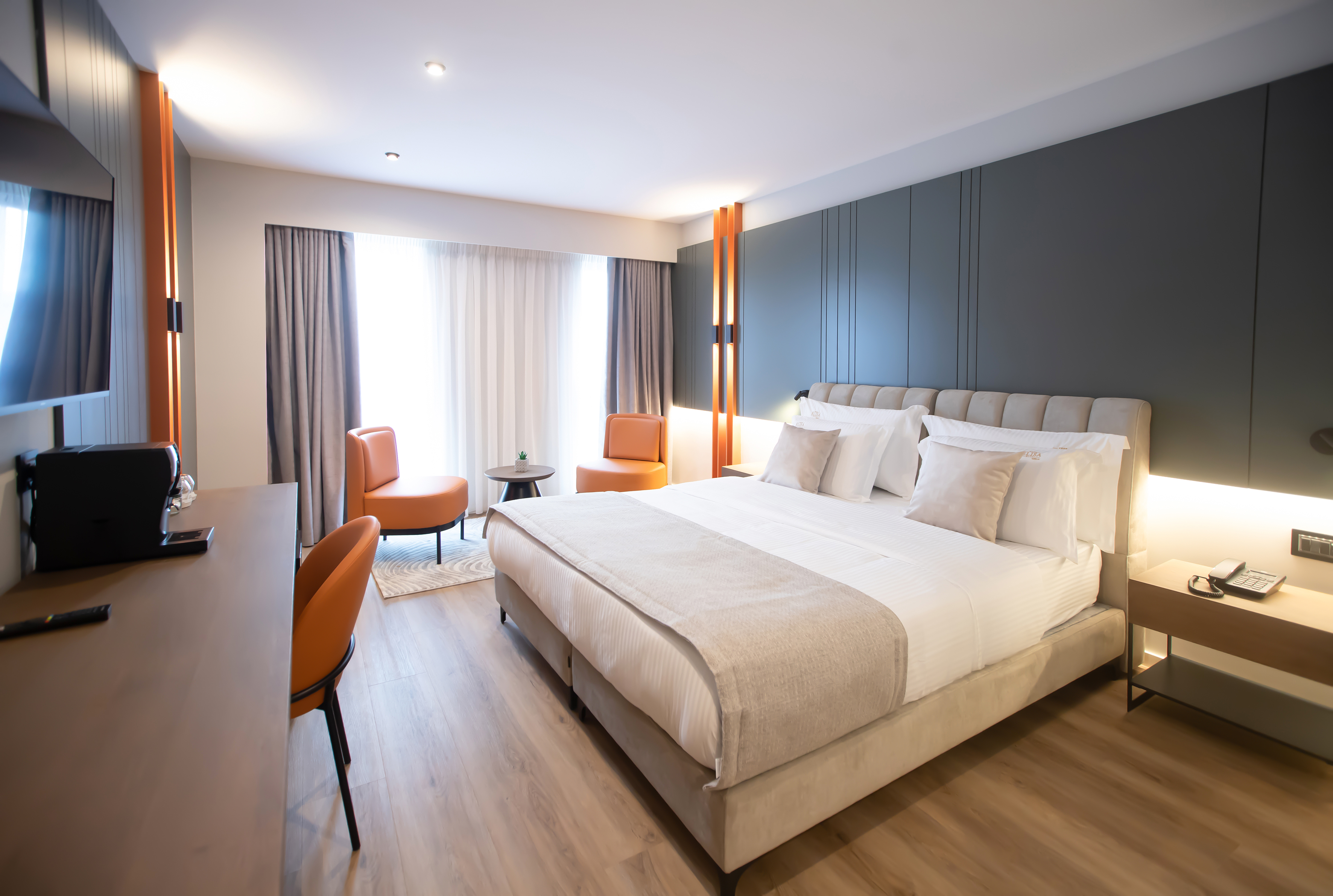 Modern hotel room with king bed, orange accent chairs, desk, and grey decor.