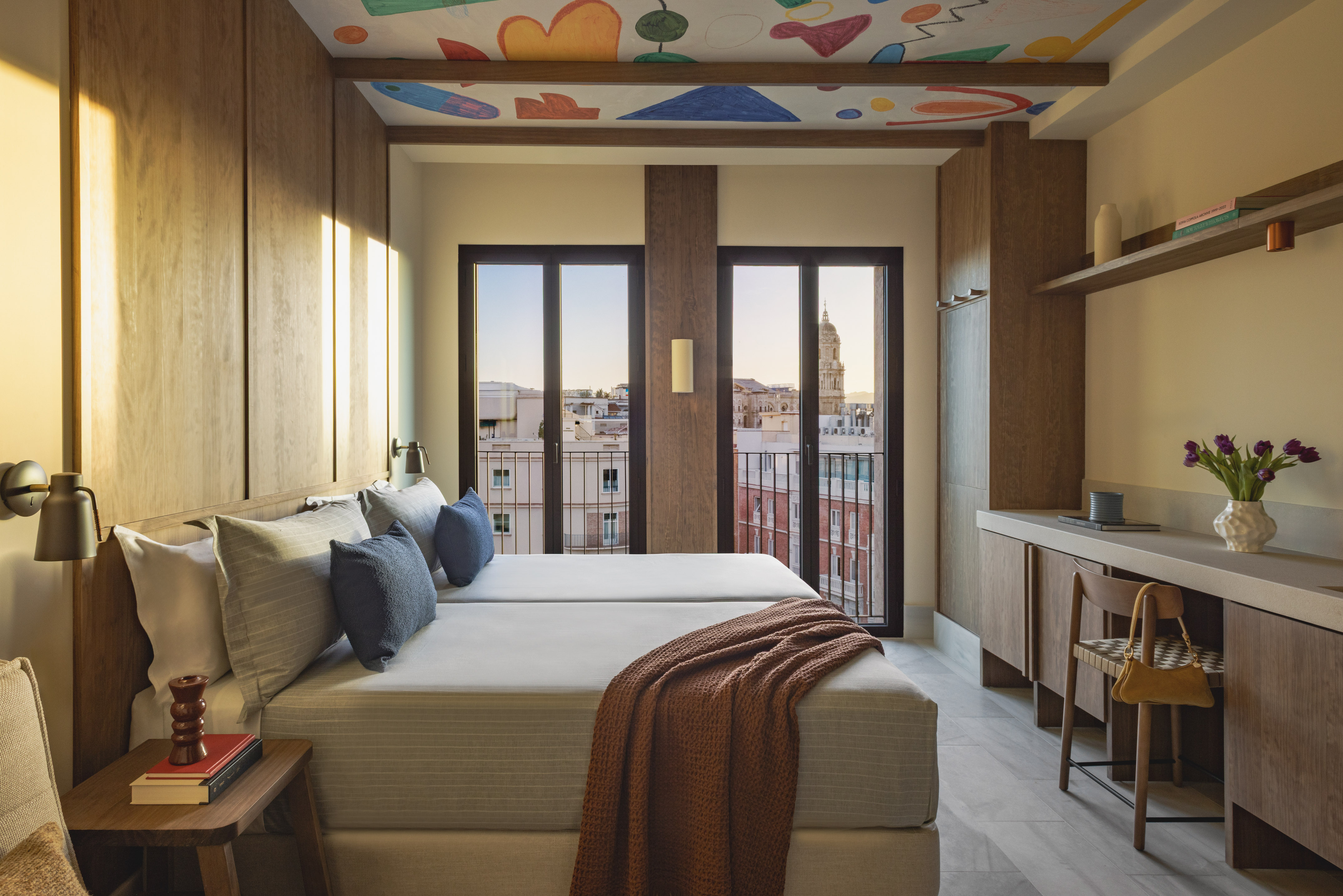 Boutique hotel room: twin beds, Malaga city view, abstract ceiling art.